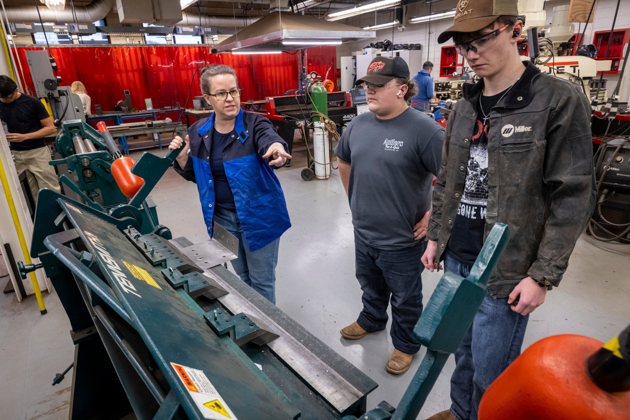 An instructor explains how to use a metal bending machine to two students in a workshop, with safety equipment and tools visible in the background.