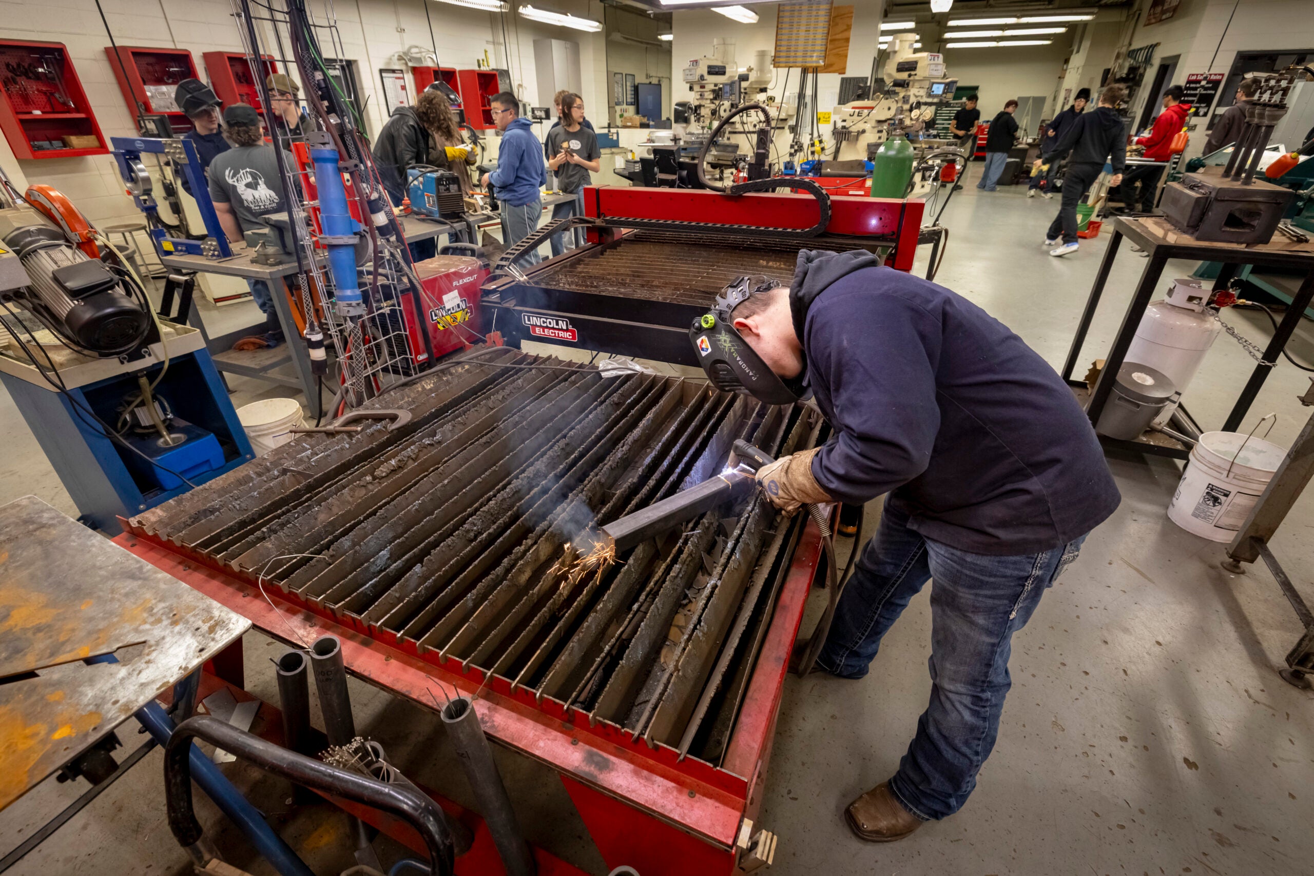 A person wearing protective gear uses a welding torch on a metal table in a busy workshop with several people and equipment in the background.