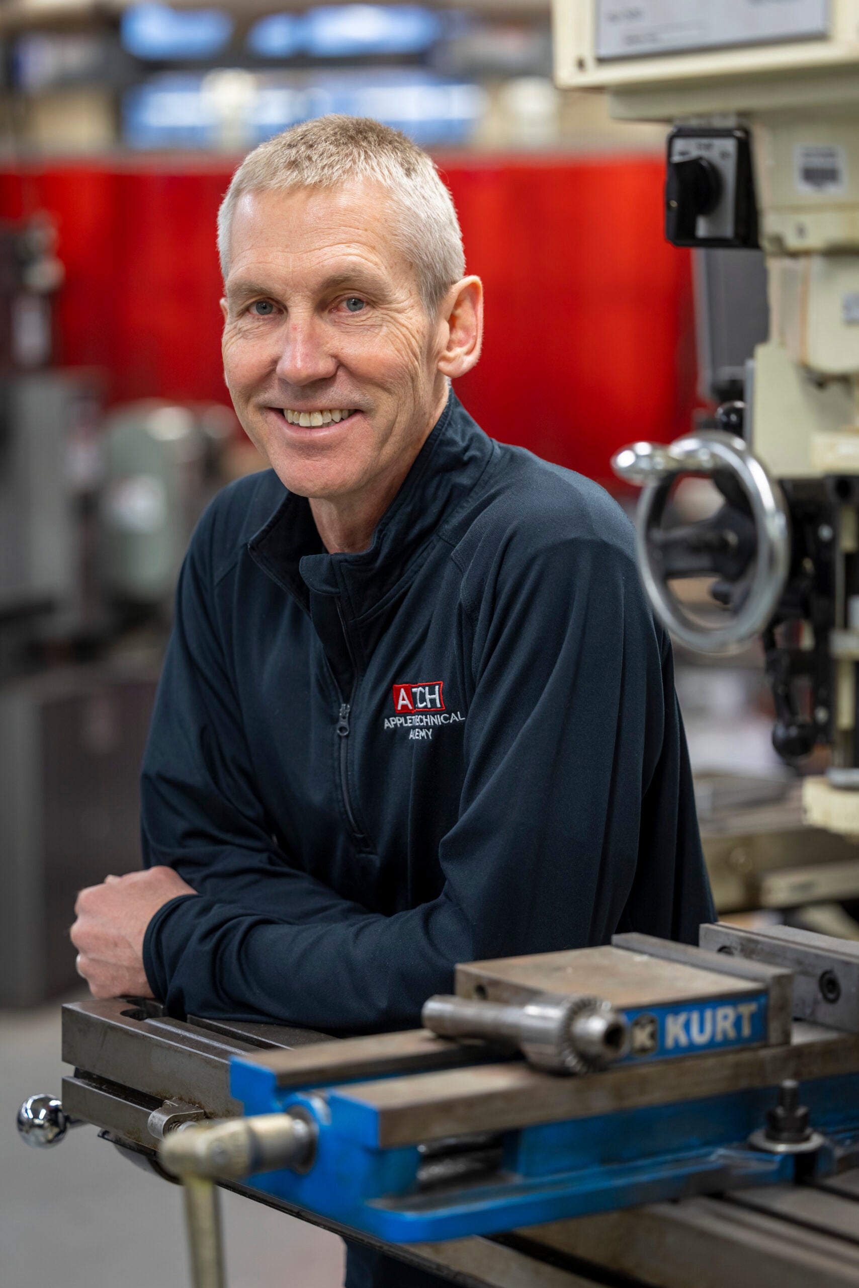 A man with short gray hair, wearing a dark jacket with an Auburn Mechanical logo, leans on a metal workbench in a workshop.