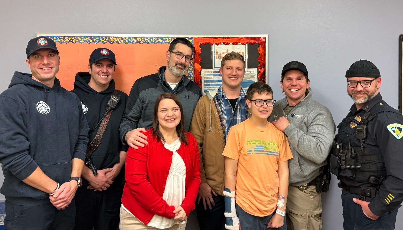 Eight people, including firefighters, a police officer, and civilians, smile while posing together indoors in front of a bulletin board.