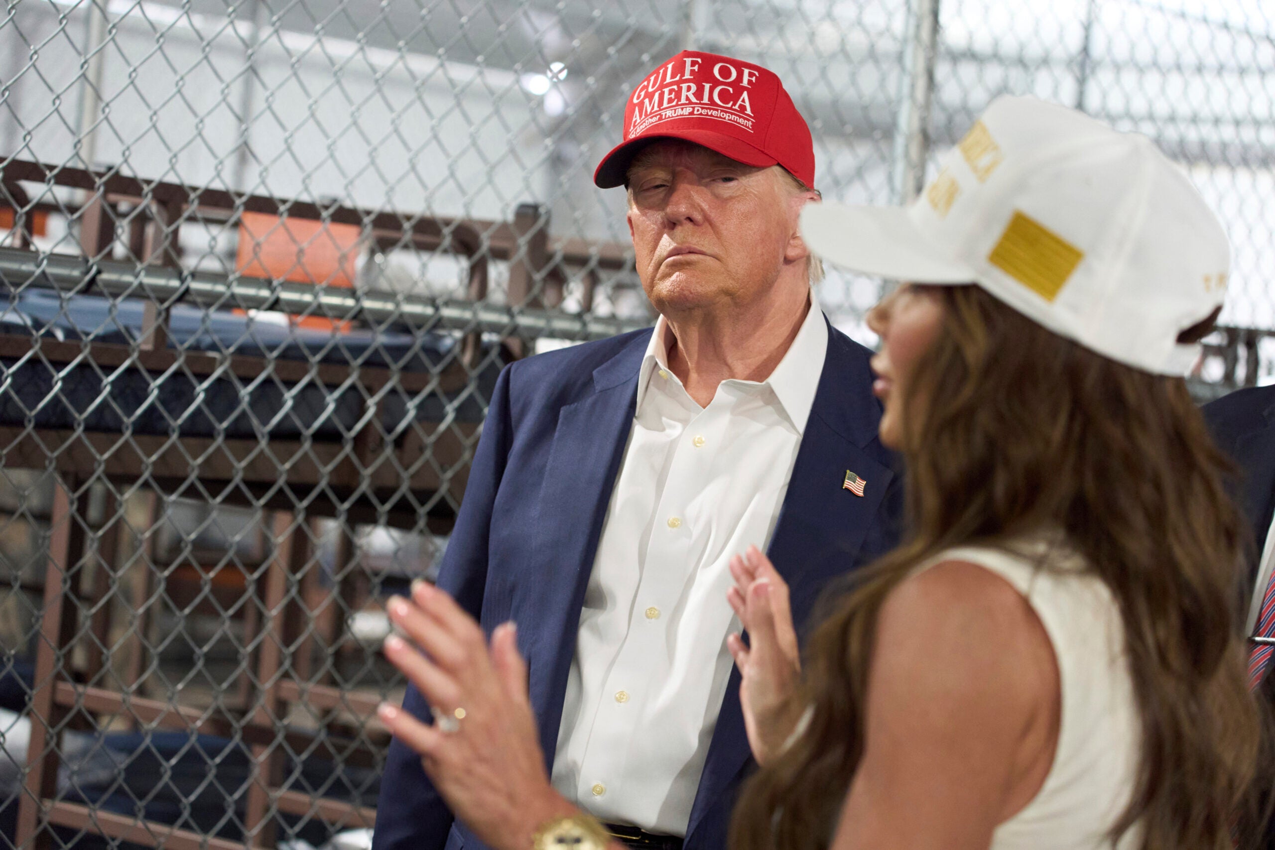 A man wearing a red Golf of America hat and suit stands near a woman in a white hat, inside a fenced indoor area with metal beds in the background.
