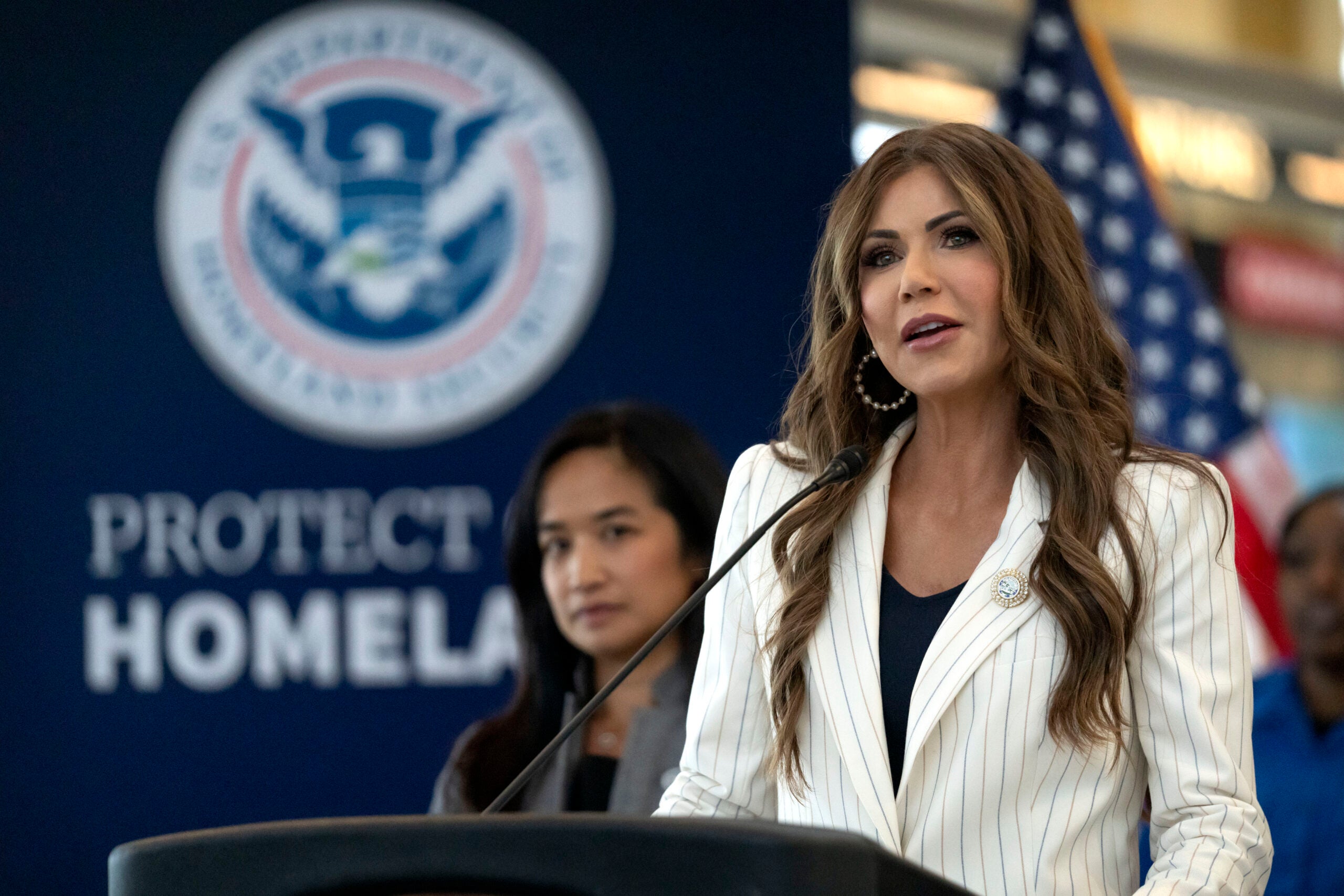 A woman speaks at a podium with a Protect Homeland sign and the U.S. Department of Homeland Security seal in the background. An American flag and another woman are also visible.