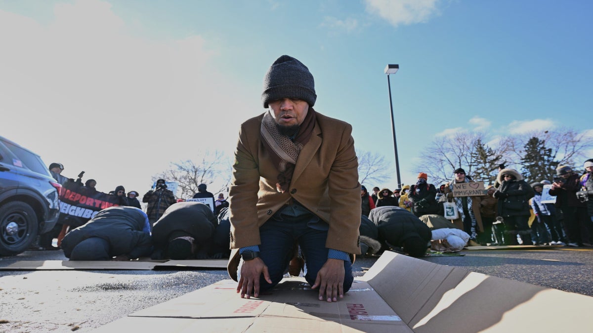 A man kneels in prayer on a cardboard sheet outdoors, while a group of people and protest signs are visible in the background.