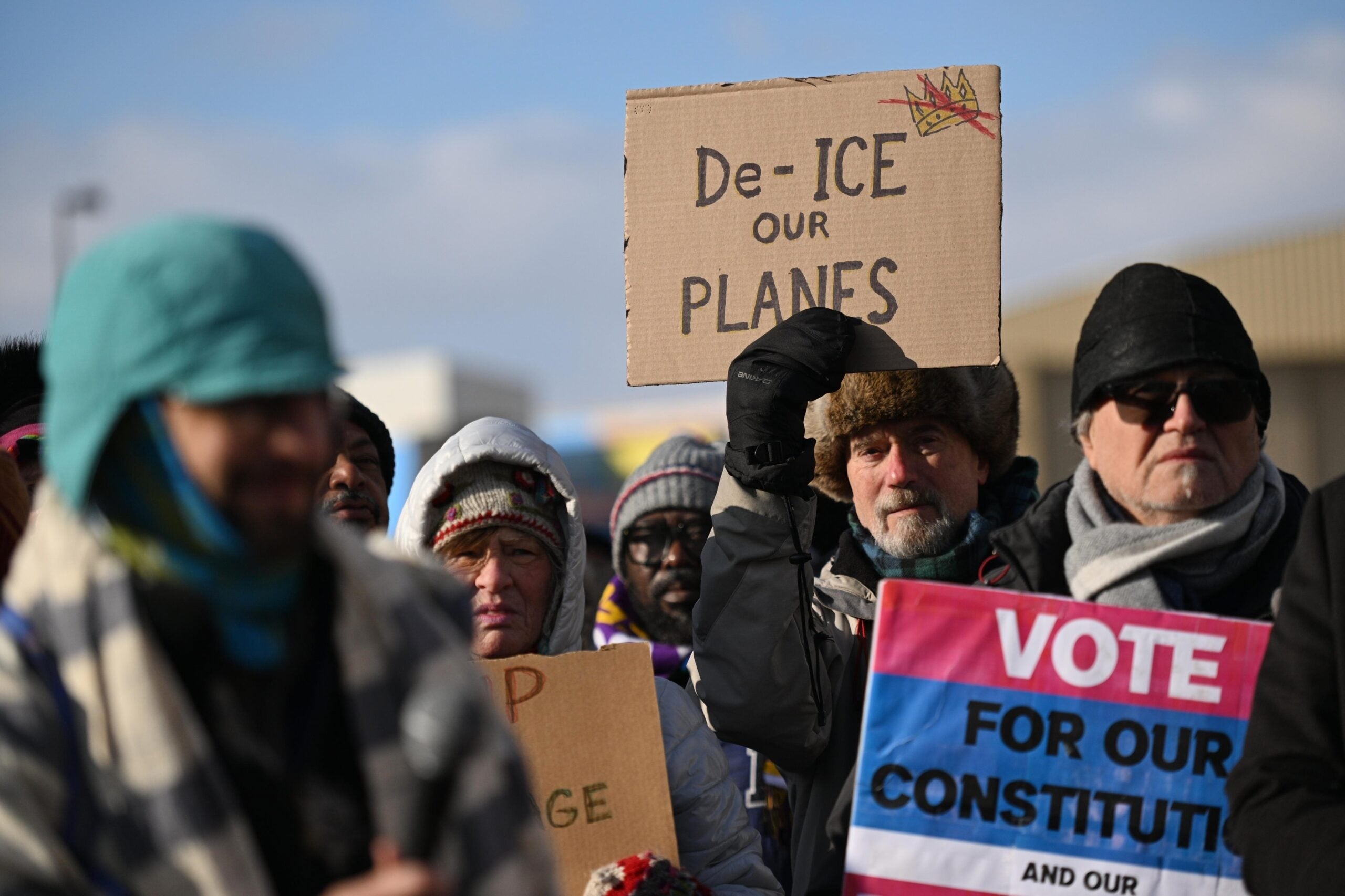 A group of bundled-up people protest outdoors; one person holds a sign reading De-ICE our PLANES while another holds a sign urging to VOTE FOR OUR CONSTITUTION.
