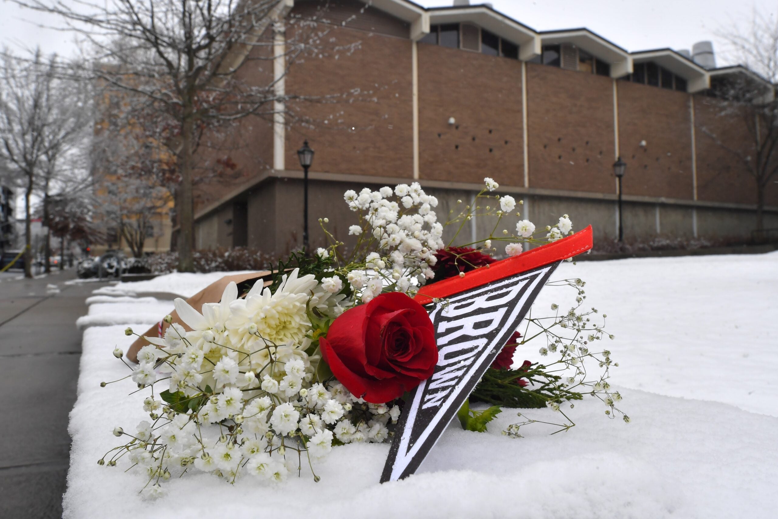A bouquet of flowers and a Brown University pennant rest on snow outside a brick building on a winter day.