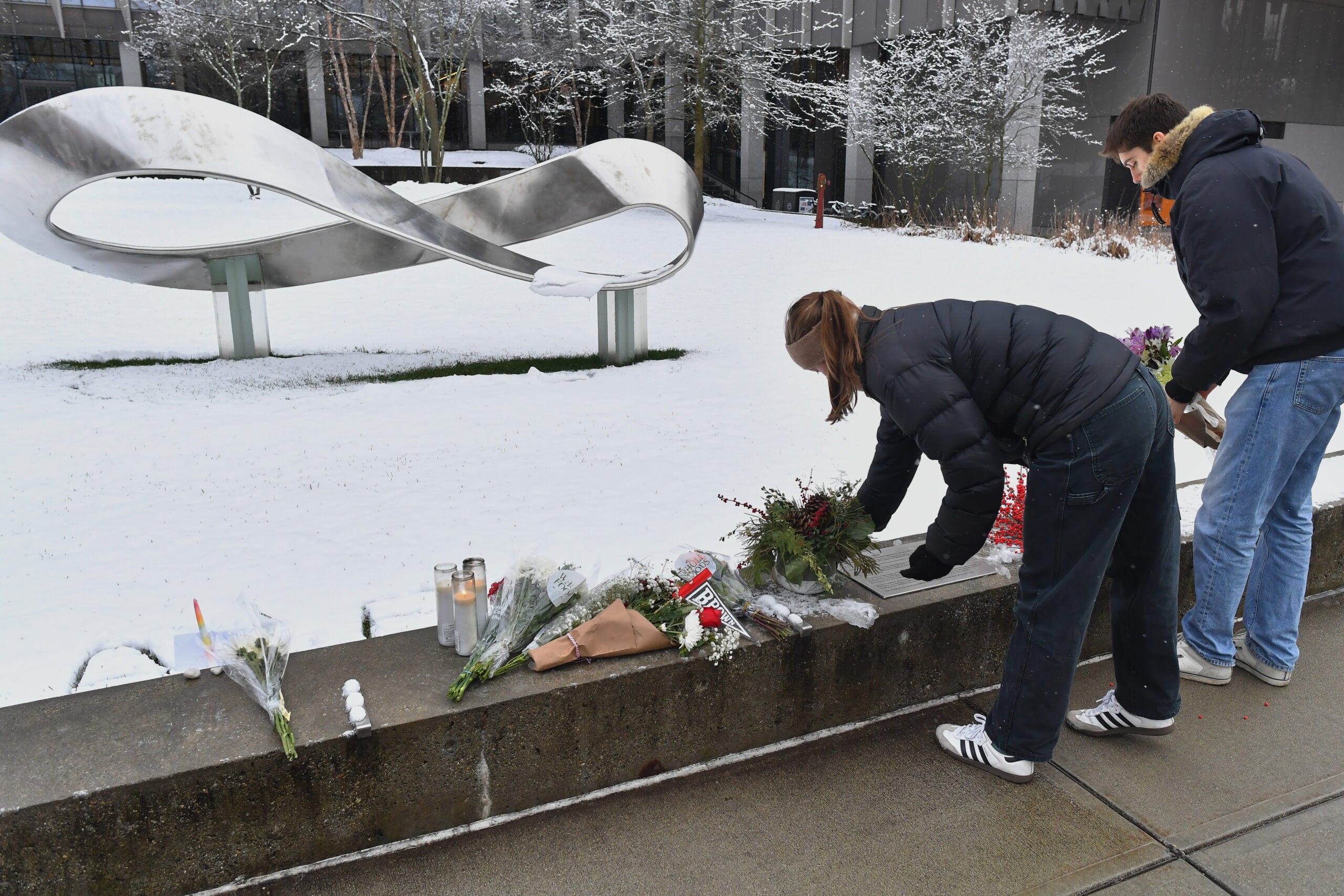 Two people place flowers and candles at a snowy outdoor memorial in front of an abstract infinity-shaped sculpture.