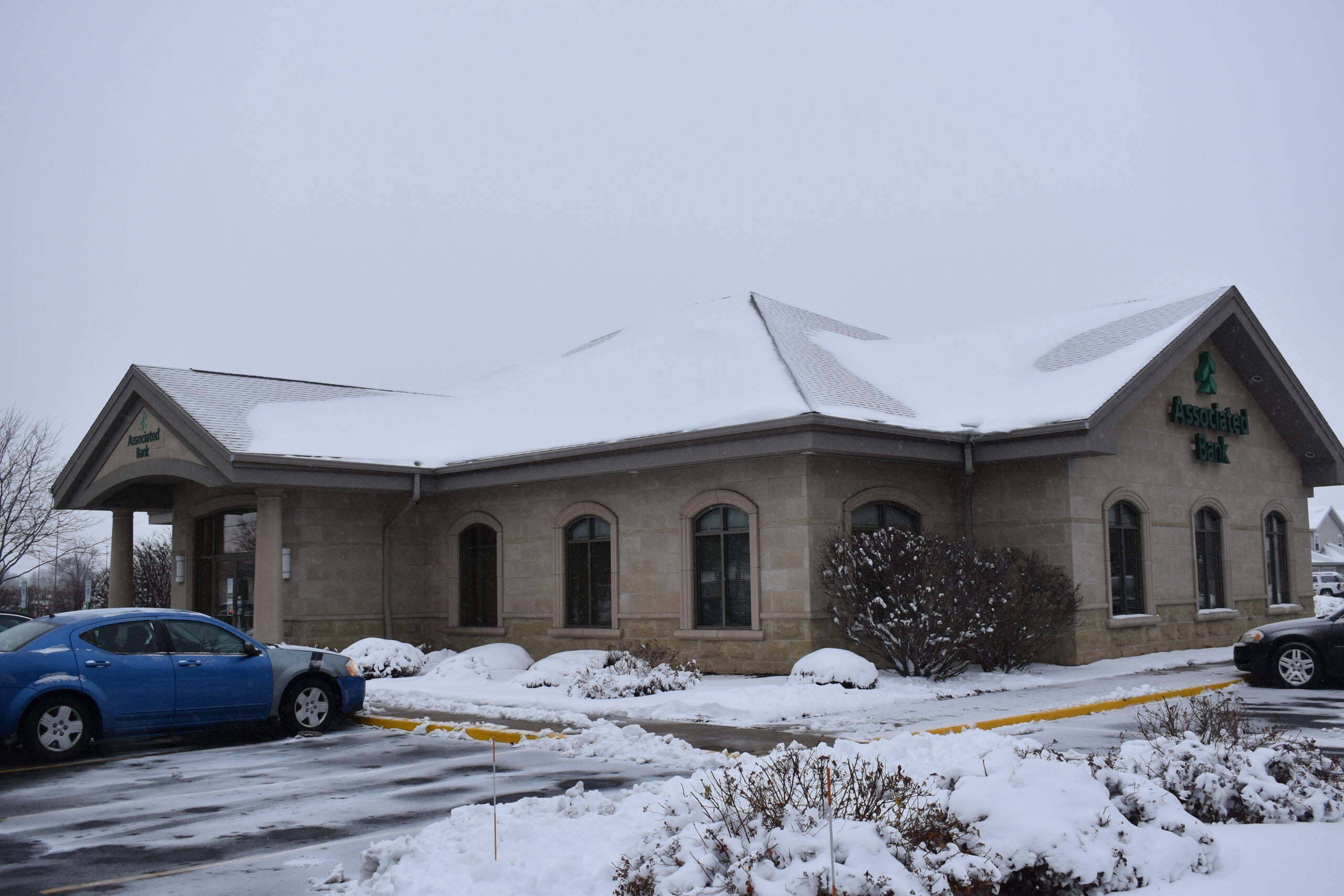 A beige stone bank building with a snow-covered roof and surrounding bushes, with two cars parked in front on a snowy, overcast day.