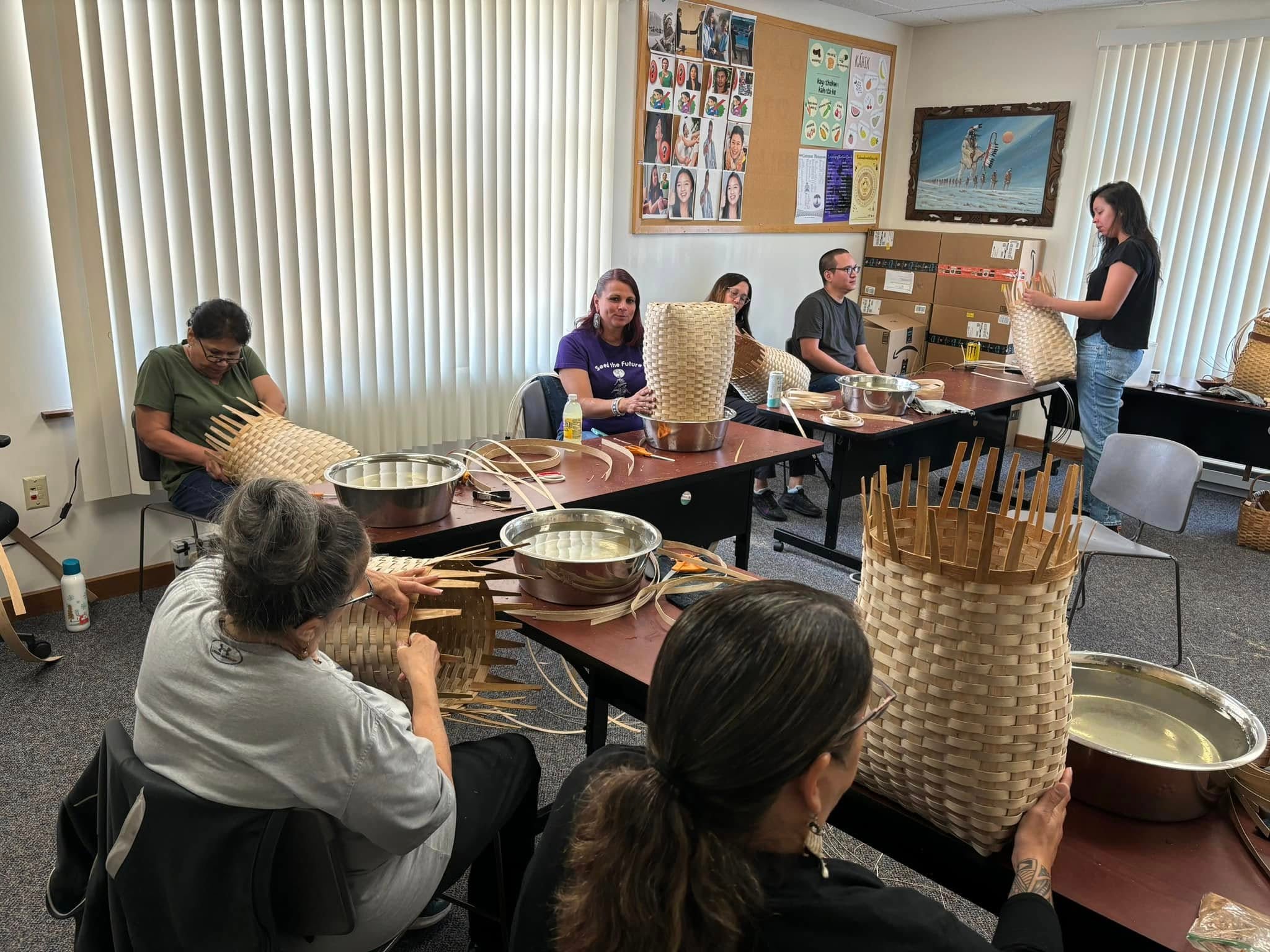 A group of people sit around tables weaving baskets in a classroom, while a woman stands at the front giving instructions.