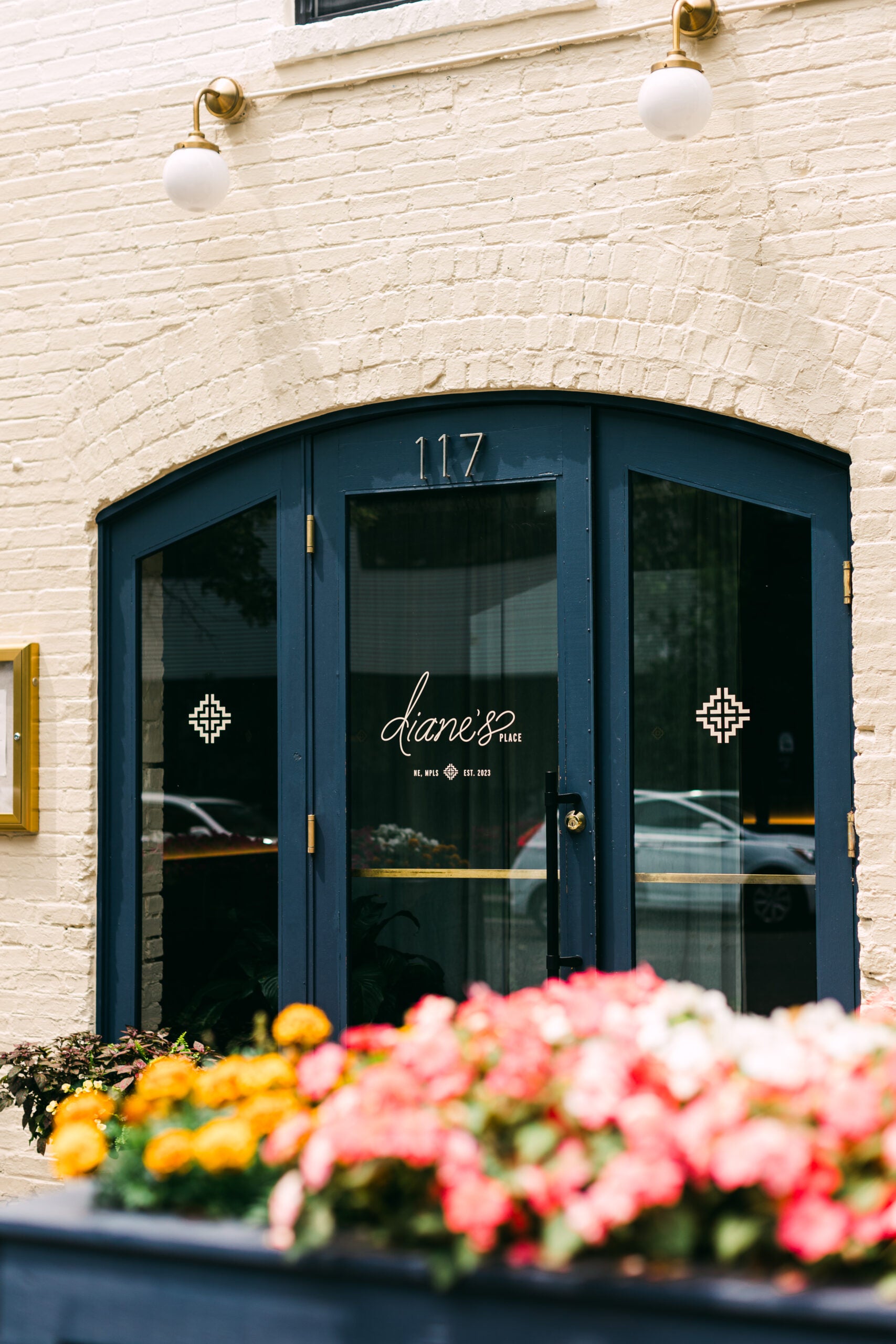 Blue double doors with Diane’s Place written on glass, set in a cream brick wall with the number 117 above; colorful flowers in the foreground.
