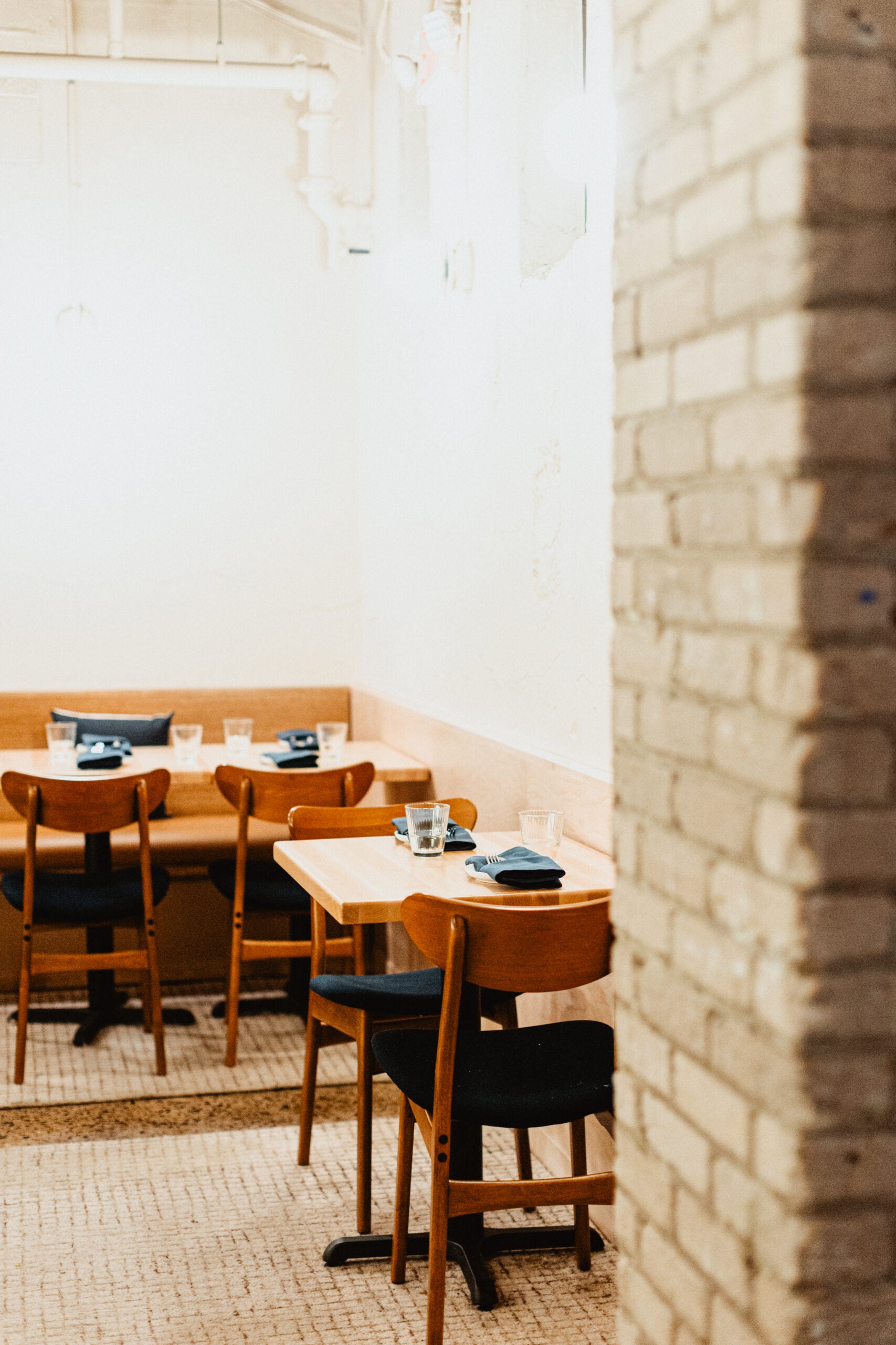A small restaurant dining area with wooden tables and chairs, blue napkins, and empty glasses, partially obscured by a brick wall in the foreground.