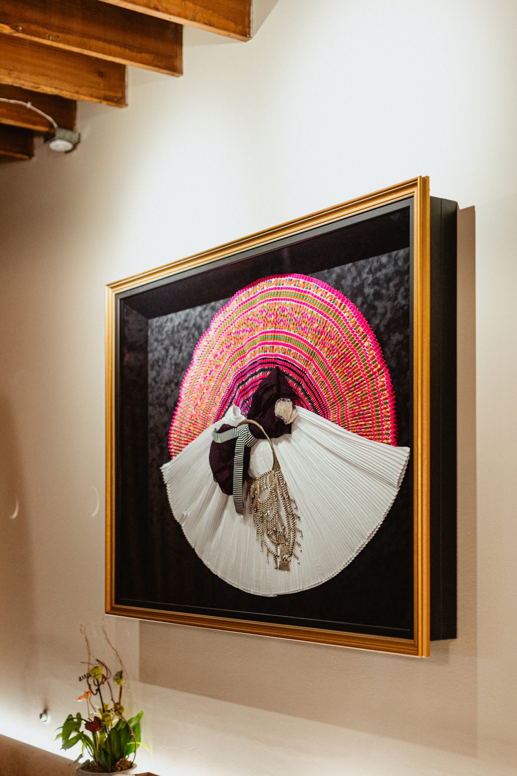 Framed artwork featuring a three-dimensional figure wearing a white dress and colorful circular headdress, mounted on a white wall with a wooden ceiling above.