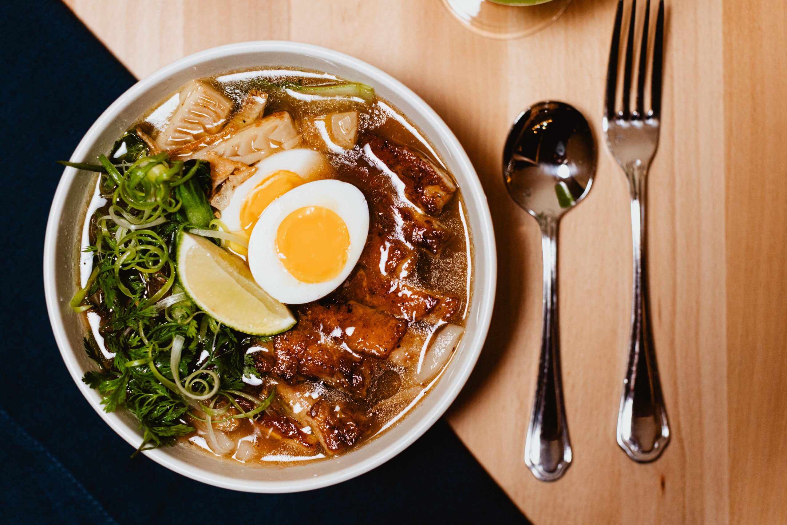 A bowl of noodle soup topped with sliced egg, lime wedge, grilled meat, herbs, green onions, and bamboo shoots, next to a spoon and fork on a wooden table.