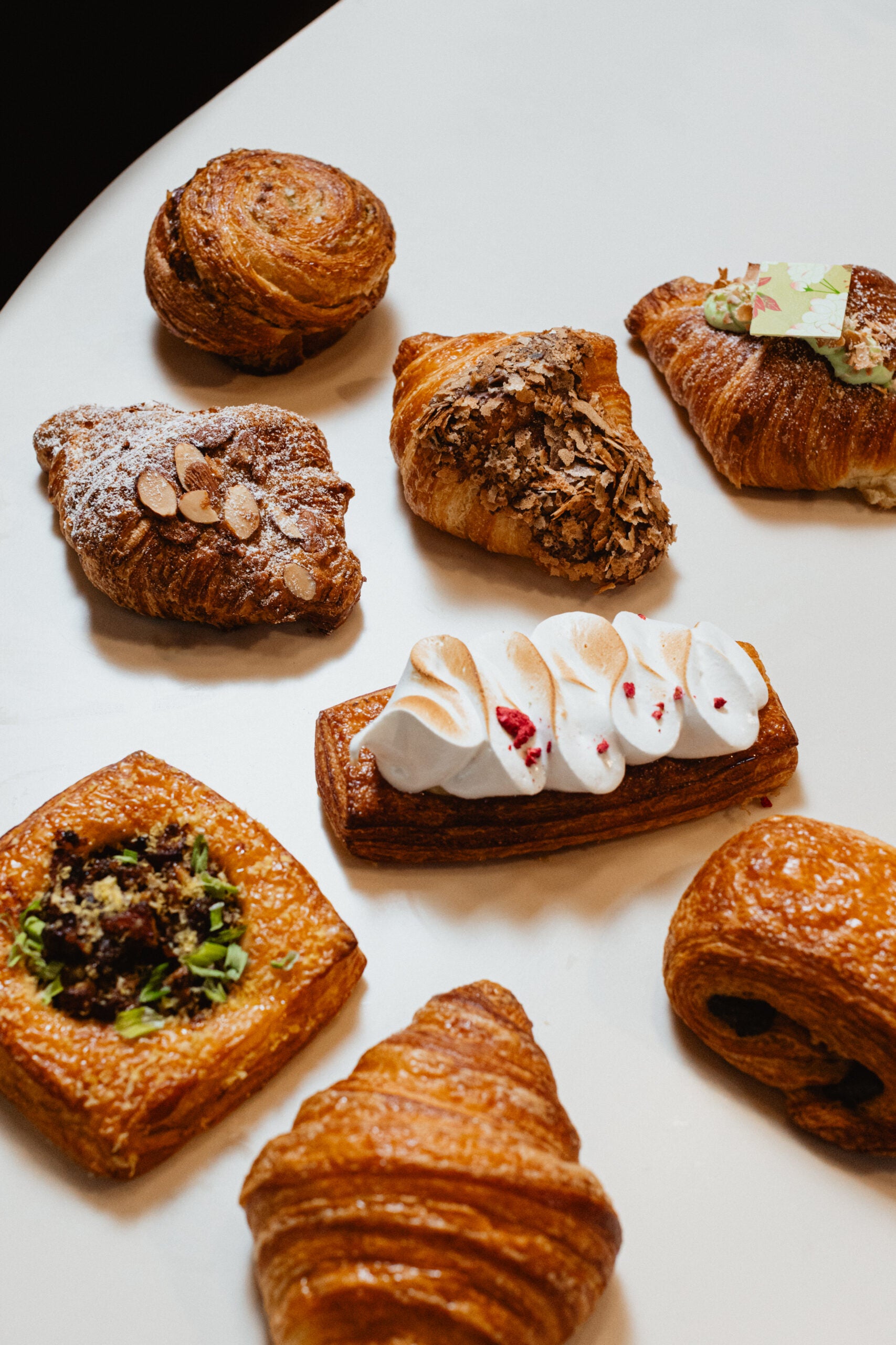 An assortment of pastries, including croissants, danishes, and a rectangular pastry topped with meringue, arranged on a white surface.