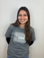 A woman with long brown hair stands against a plain white wall, smiling and wearing a gray t-shirt over a black long-sleeve shirt.