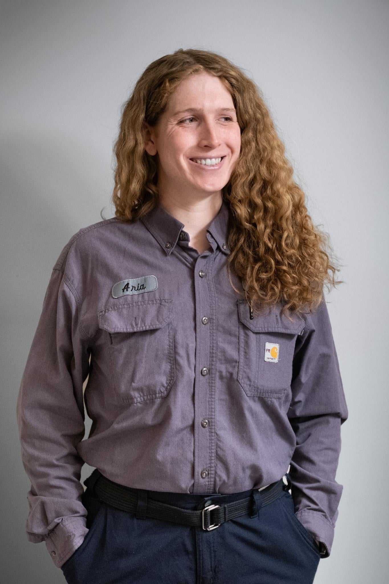 A person with long curly hair, wearing a gray button-up shirt with a name tag that says Anita, stands against a plain background and smiles while looking to the side.