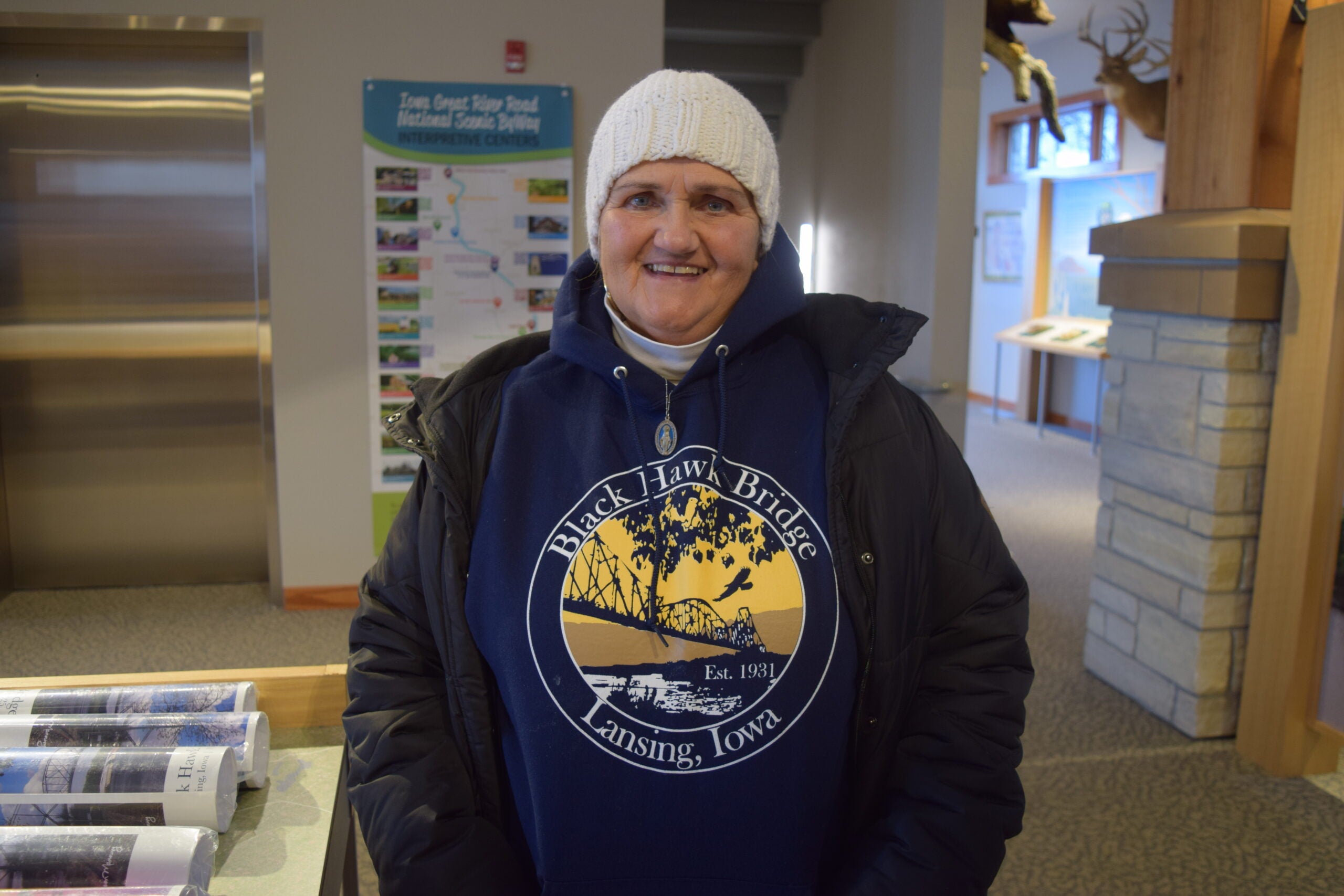 A woman wearing a white knit hat and a Black Hawk Bridge Lansing, Iowa hoodie stands indoors, smiling at the camera.