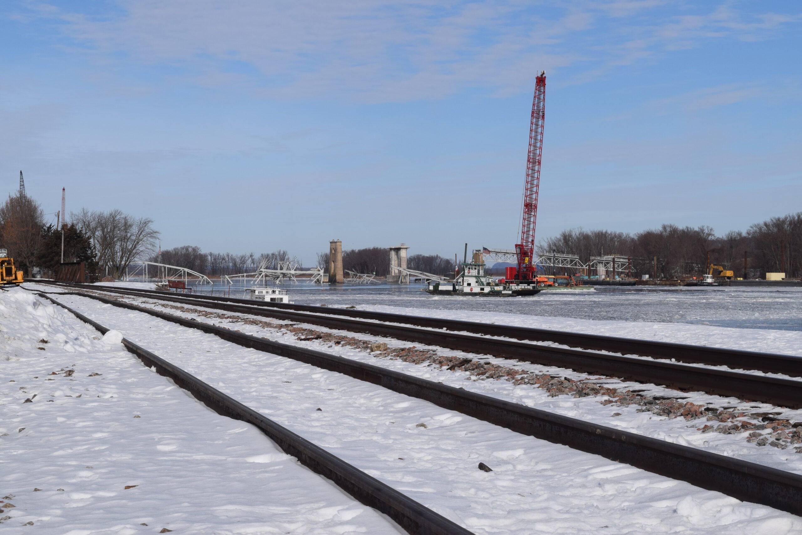 Snow-covered railway tracks run alongside a river, with a red crane, a barge, and a partially raised drawbridge visible in the background under a partly cloudy sky.