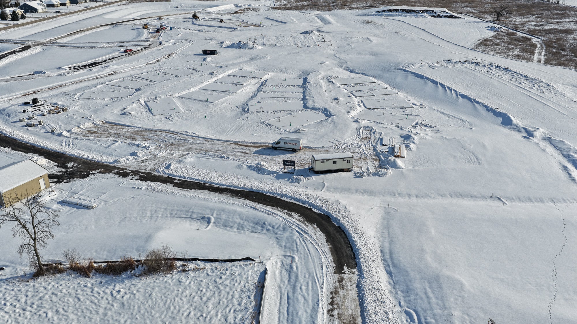 A snow-covered construction site with visible building foundations, a few vehicles, and portable office structures, surrounded by roads and open land.