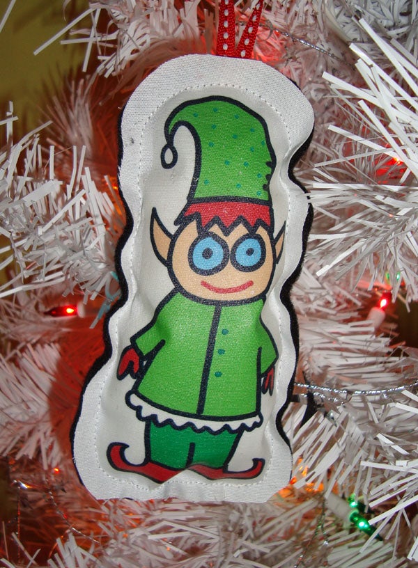 A fabric ornament featuring a cartoon elf in green clothes and hat hangs on a white artificial Christmas tree with colored lights.