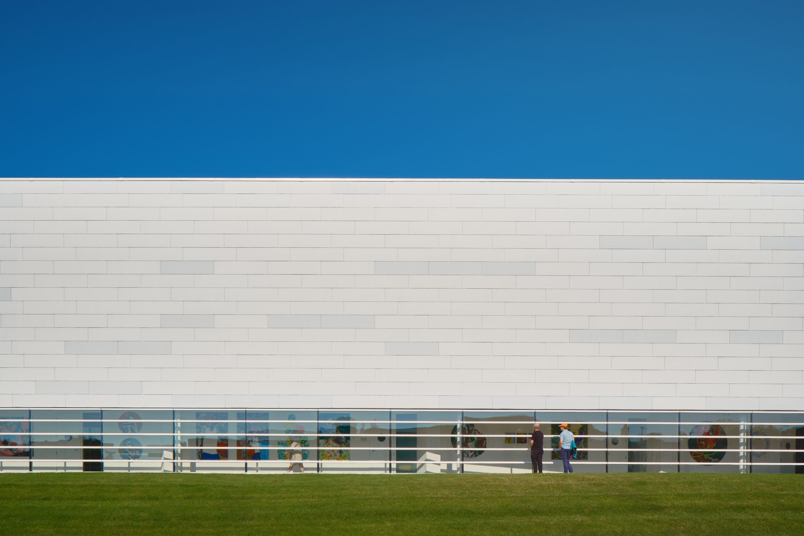 A modern white building with a flat roof and rectangular windows; three people stand outside near the windows on a sunny day with a clear blue sky.