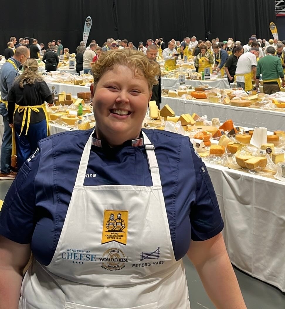 A person wearing a white apron and navy shirt stands smiling in front of tables covered with various cheeses at a busy event.