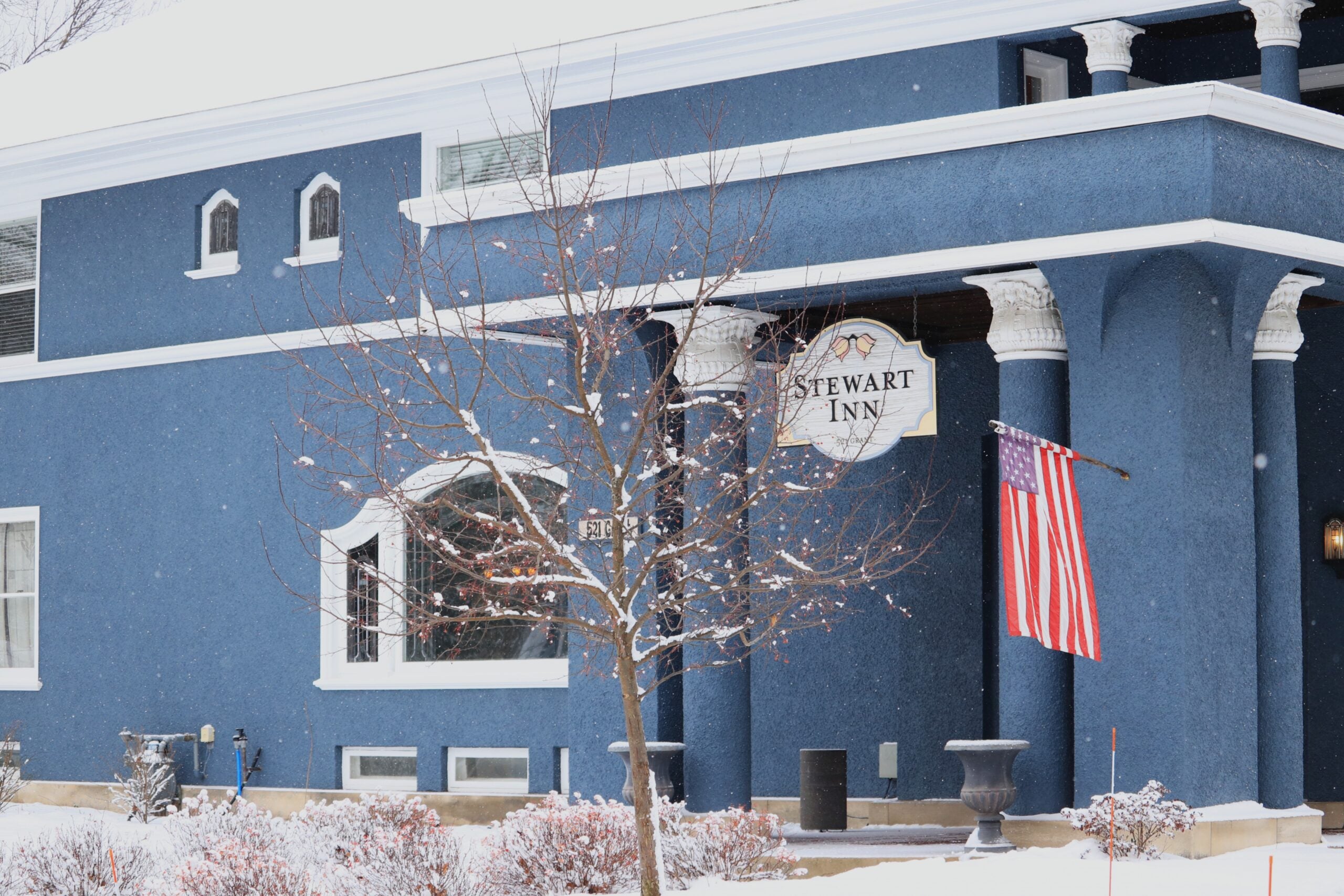 A blue building labeled Stewart Inn with white trim, an American flag, and light snow covering the ground and bare tree branches.