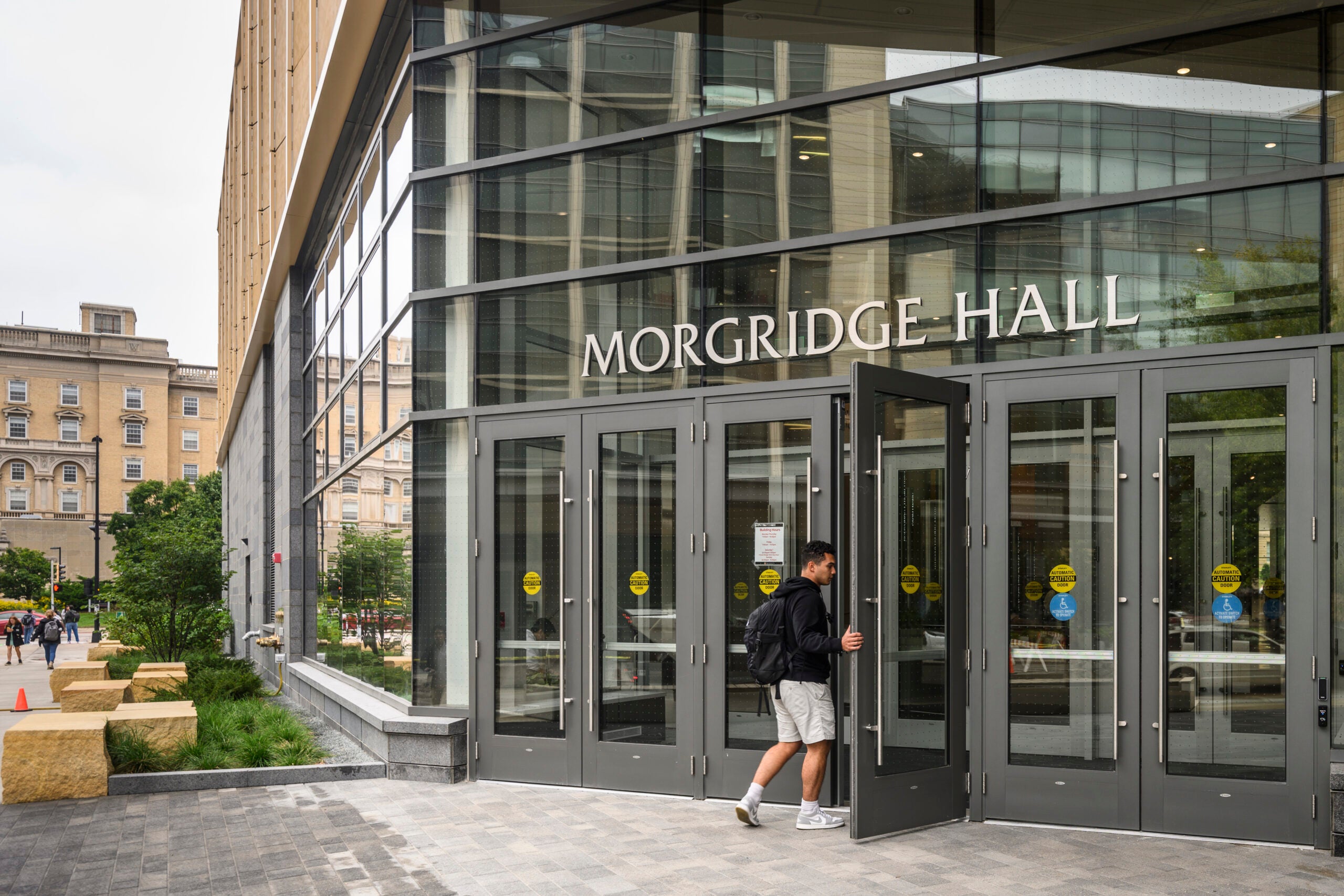 A person with a backpack enters Morgridge Hall through glass doors on a modern campus building, with older buildings visible in the background.