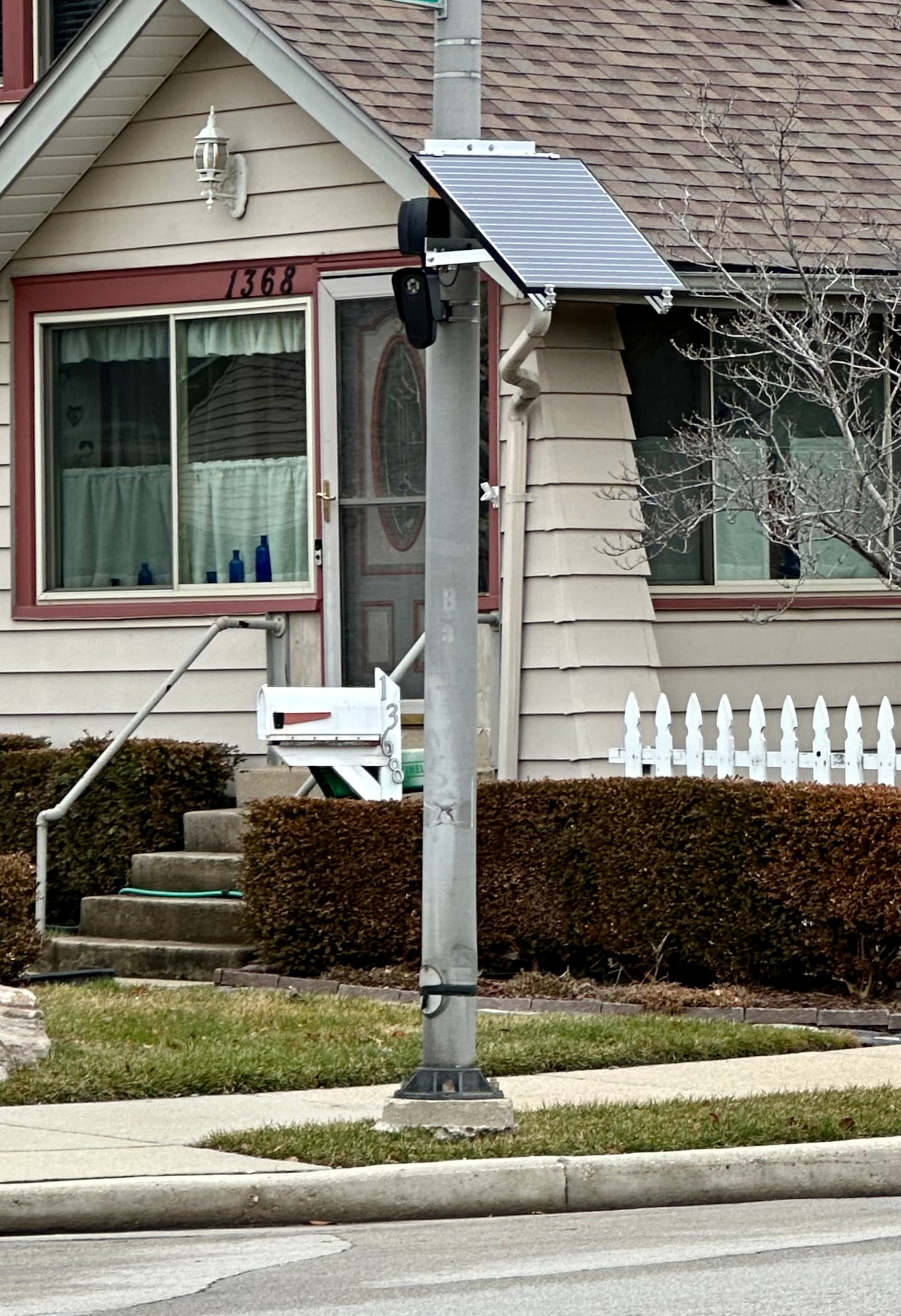 A utility pole with a solar panel and mounted camera stands near a sidewalk in front of a beige house with red trim and a manicured hedge.