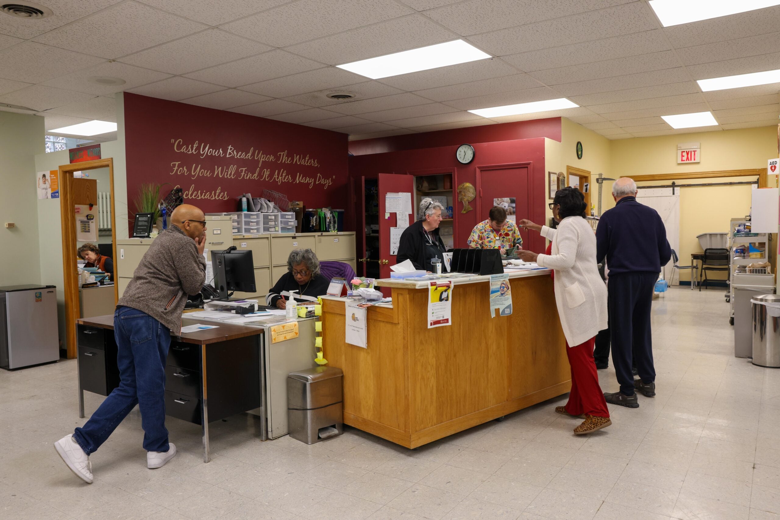 Several people interact at a reception desk in a community center or office, with staff assisting visitors and paperwork visible on the counter.