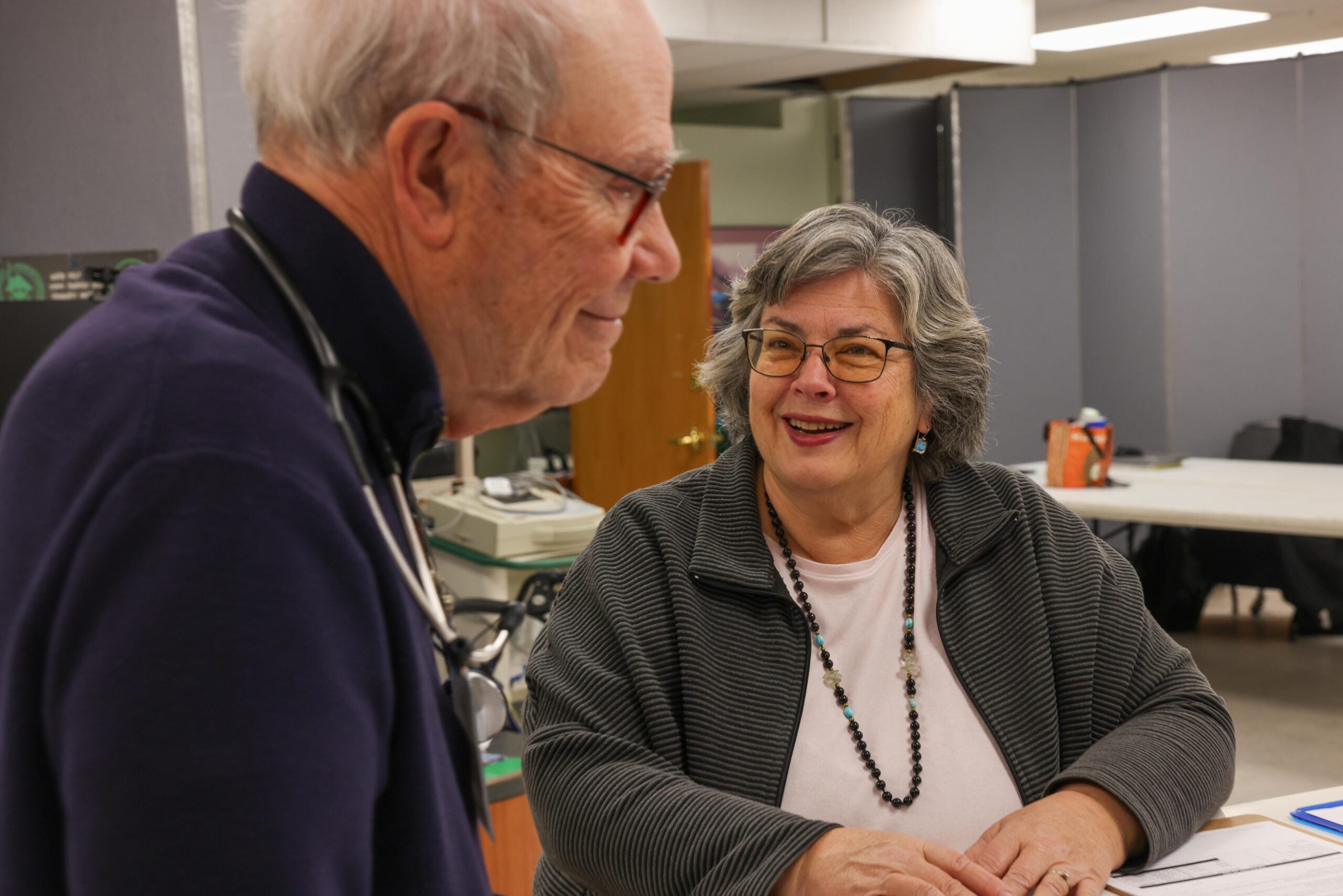 An older man and woman have a conversation at a table in an office or clinic setting; the woman is smiling and wearing glasses.