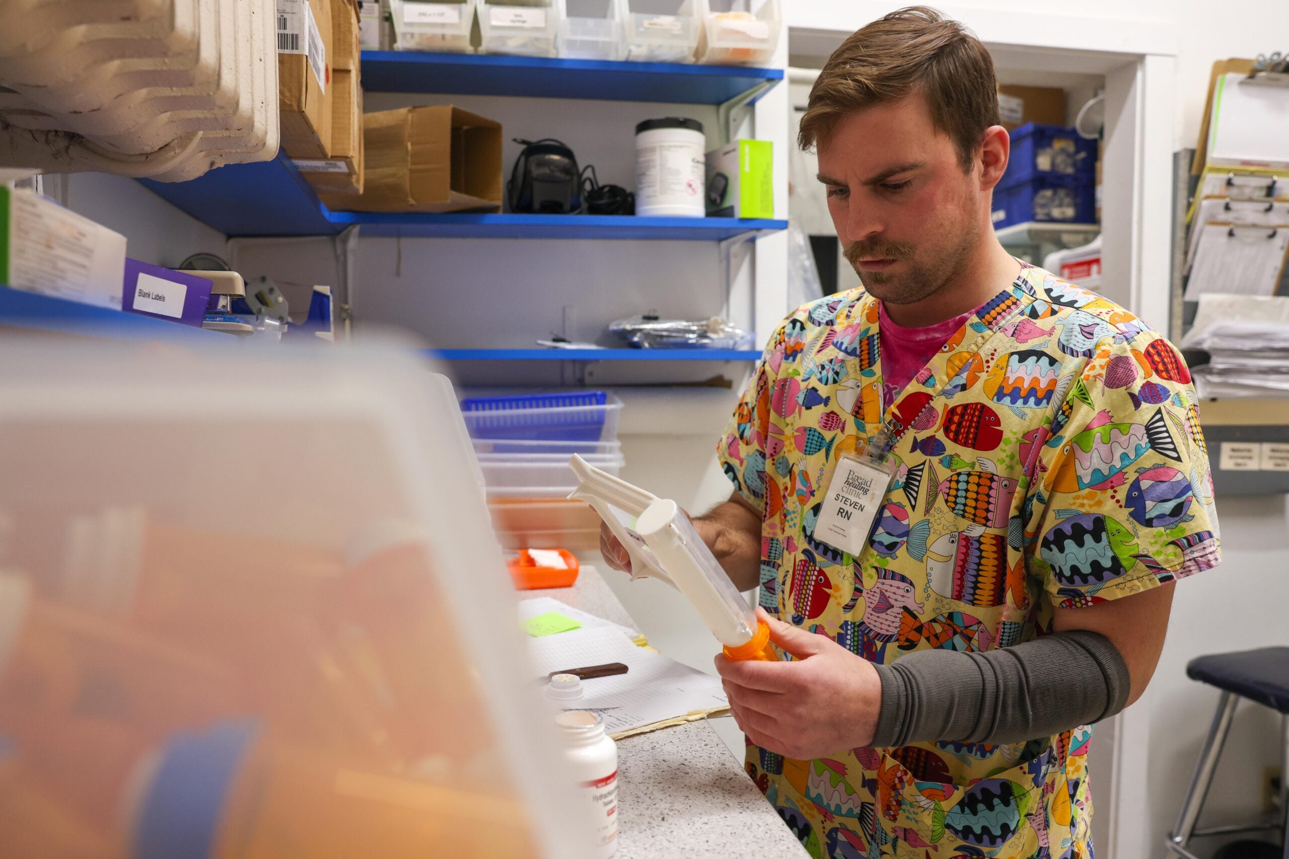 A pharmacy worker in colorful scrubs fills a prescription bottle in a pharmacy storage area with shelves holding various supplies.