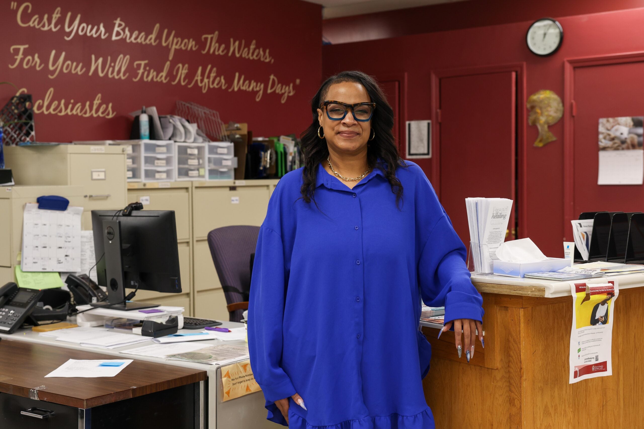 A woman in a bright blue dress stands by a wooden counter in an office with maroon walls, filing cabinets, and a quote on the wall behind her.