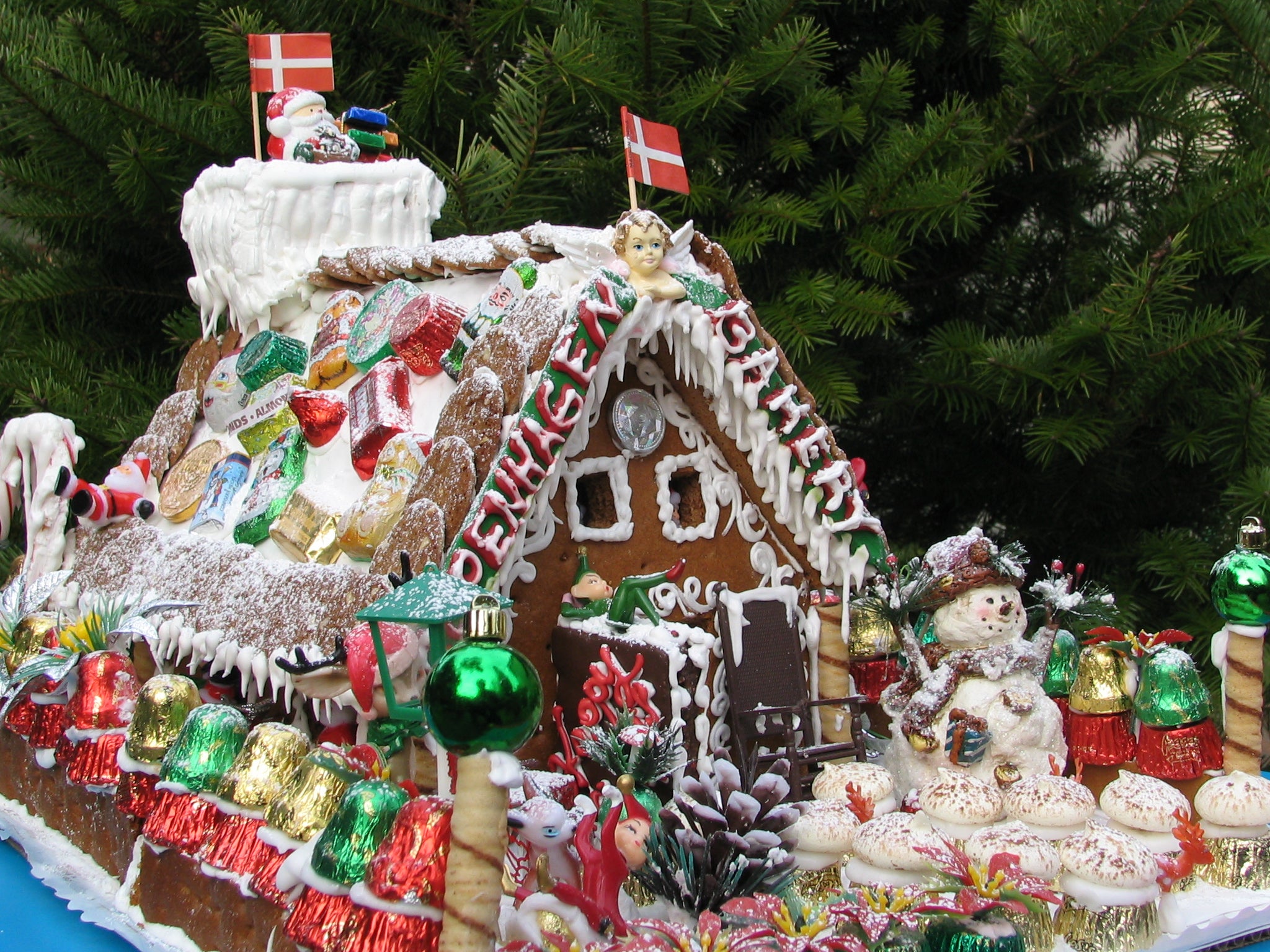 A decorated gingerbread house with Danish flags, festive candies, icing, a snowman, and Christmas ornaments in front of green pine branches.