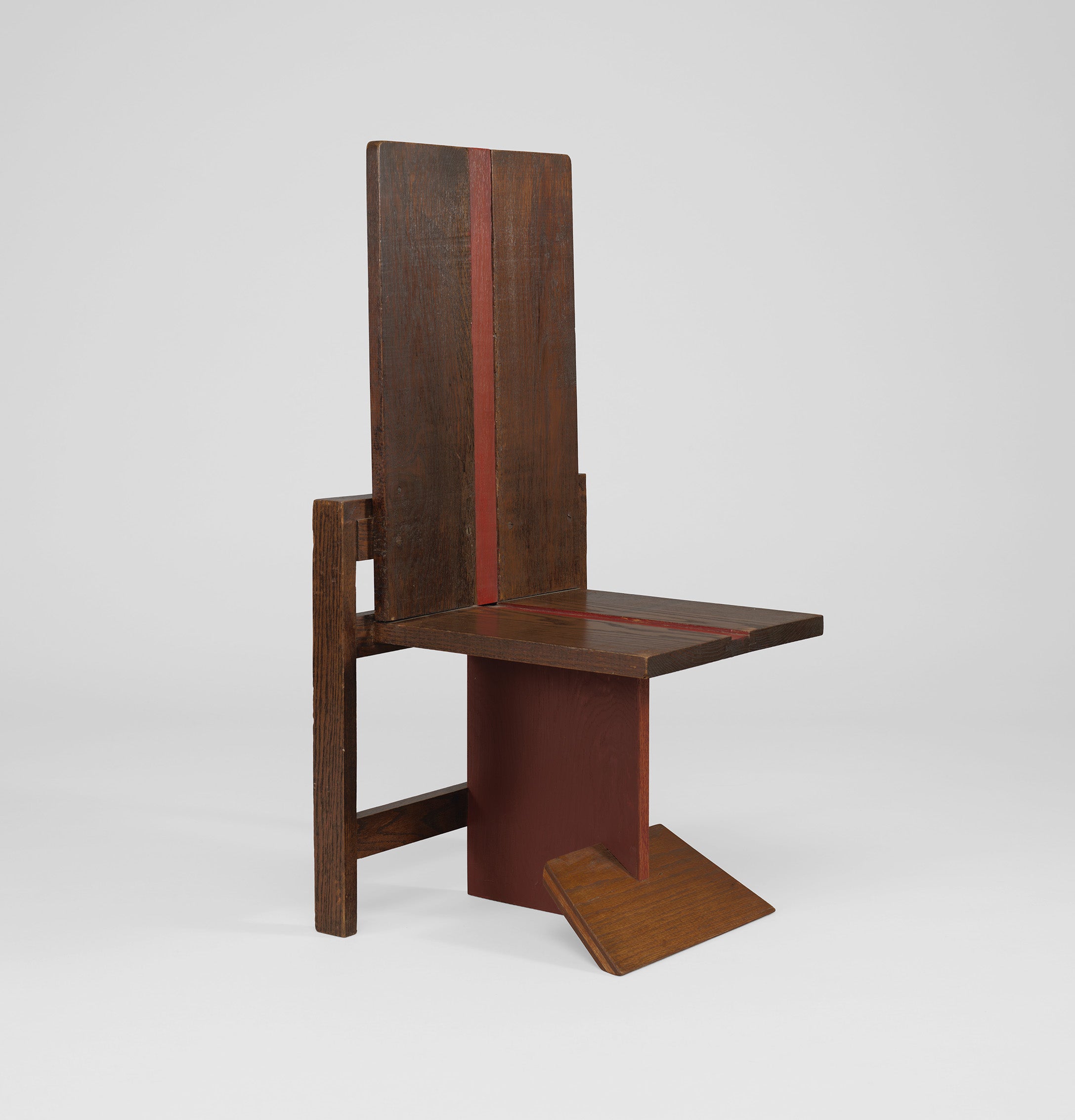 A wooden chair with a tall, flat backrest, angular seat, and geometric base featuring vertical and horizontal red-brown panels.