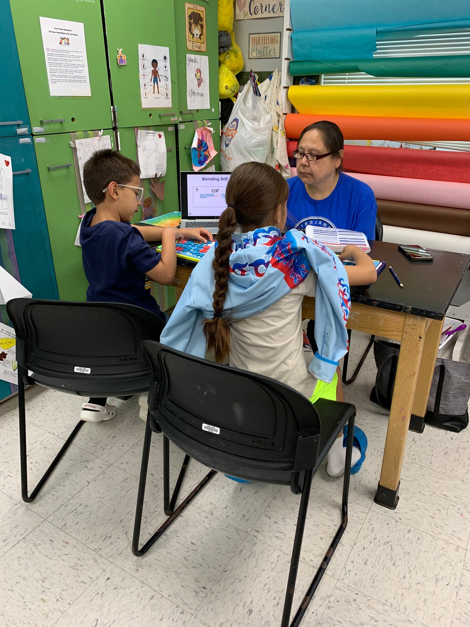 A teacher sits at a desk with two students, assisting them with schoolwork in a classroom with colorful supplies and cabinets.