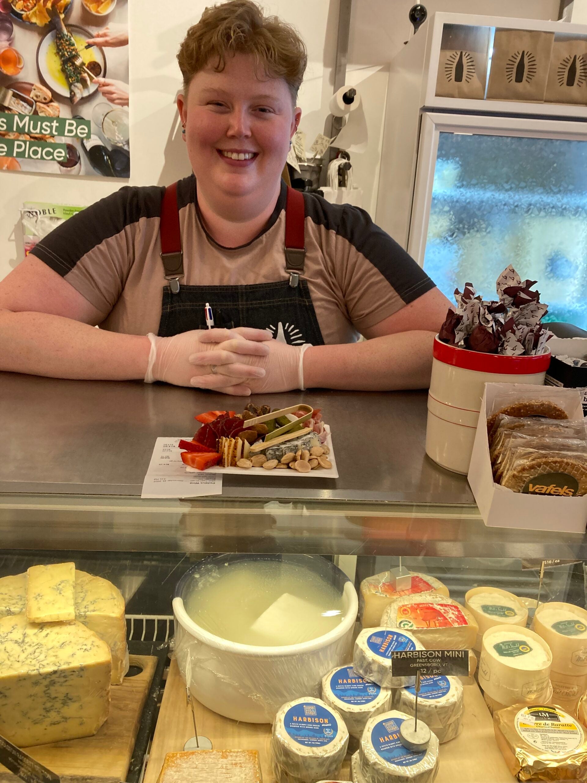 Smiling person in an apron stands behind a counter with cheeses, crackers, and a charcuterie board on display. Refrigerated cheese and dairy products are visible in the foreground.