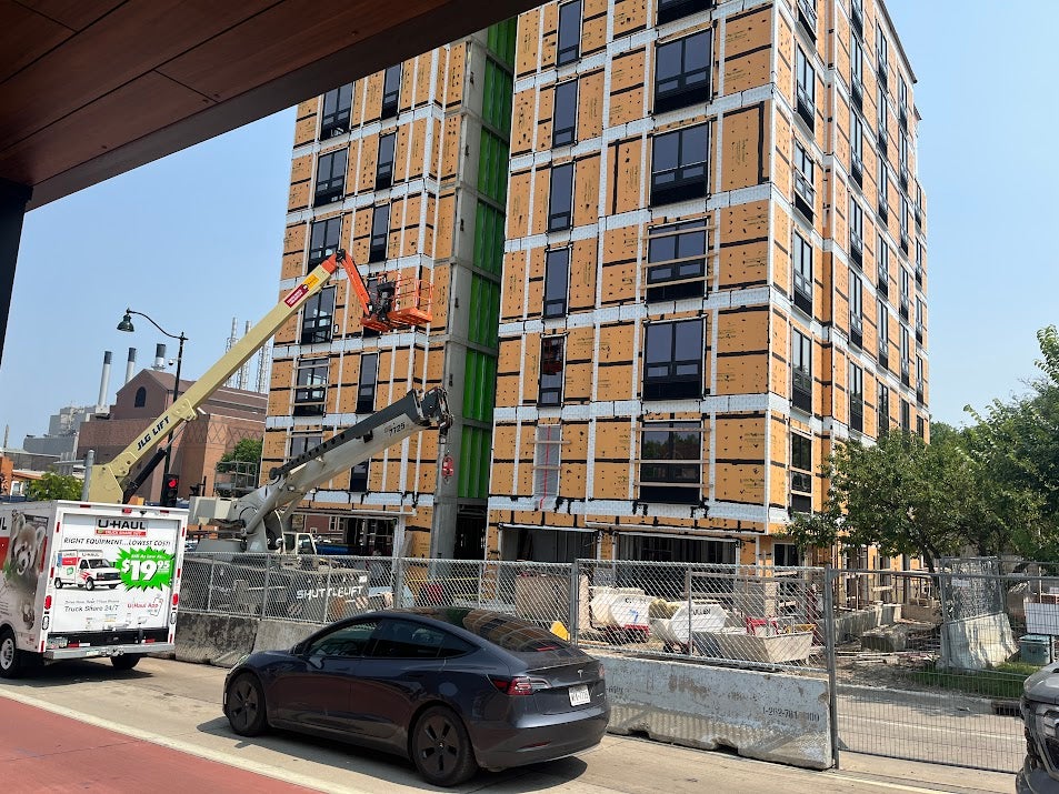 An under-construction multi-story building with exposed insulation and a crane, next to a U-Haul truck and a parked car on a city street.