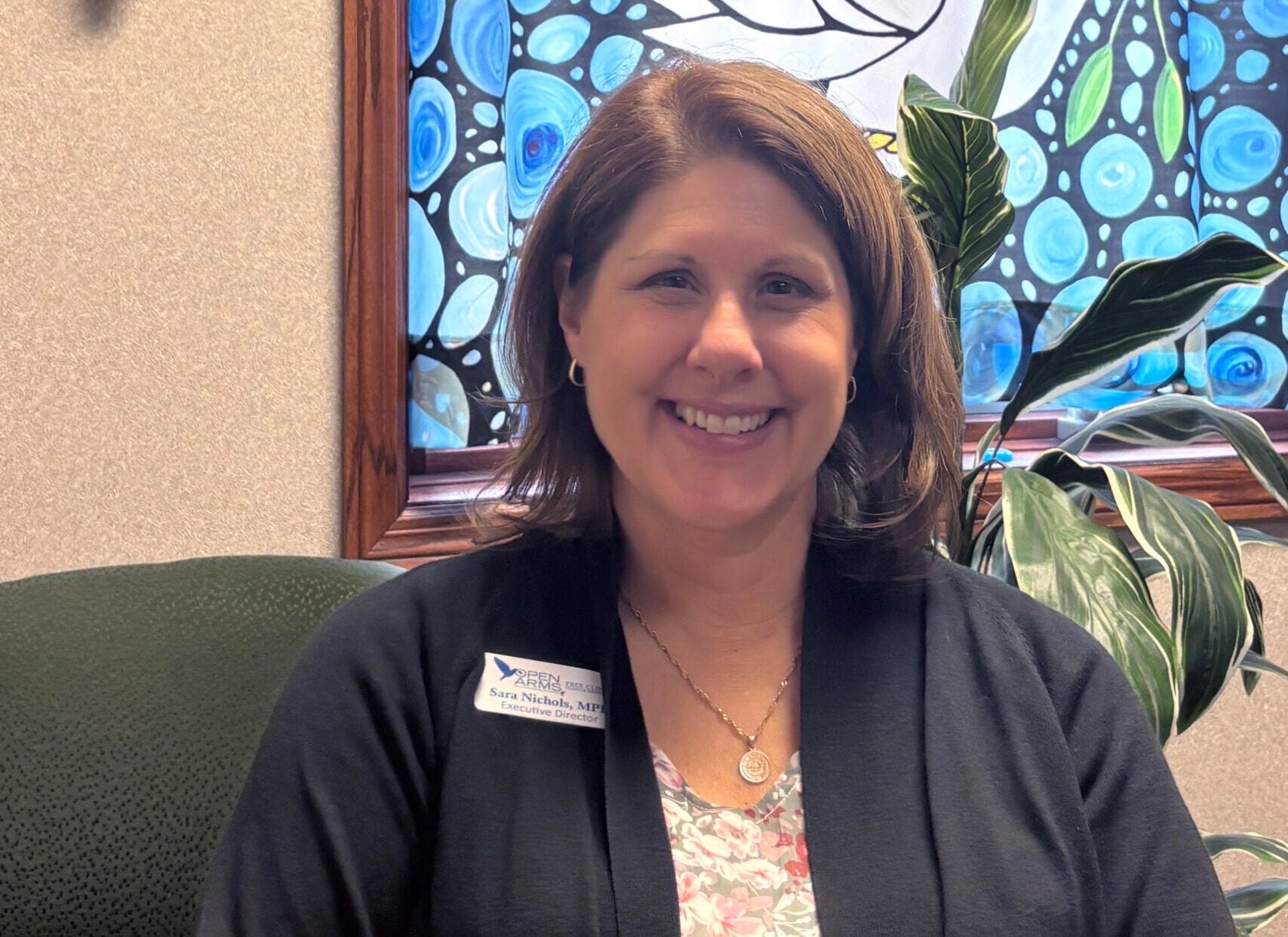 A woman sits in an office chair, smiling at the camera, wearing a black cardigan and floral top. A large artwork with a white dove is displayed behind her.