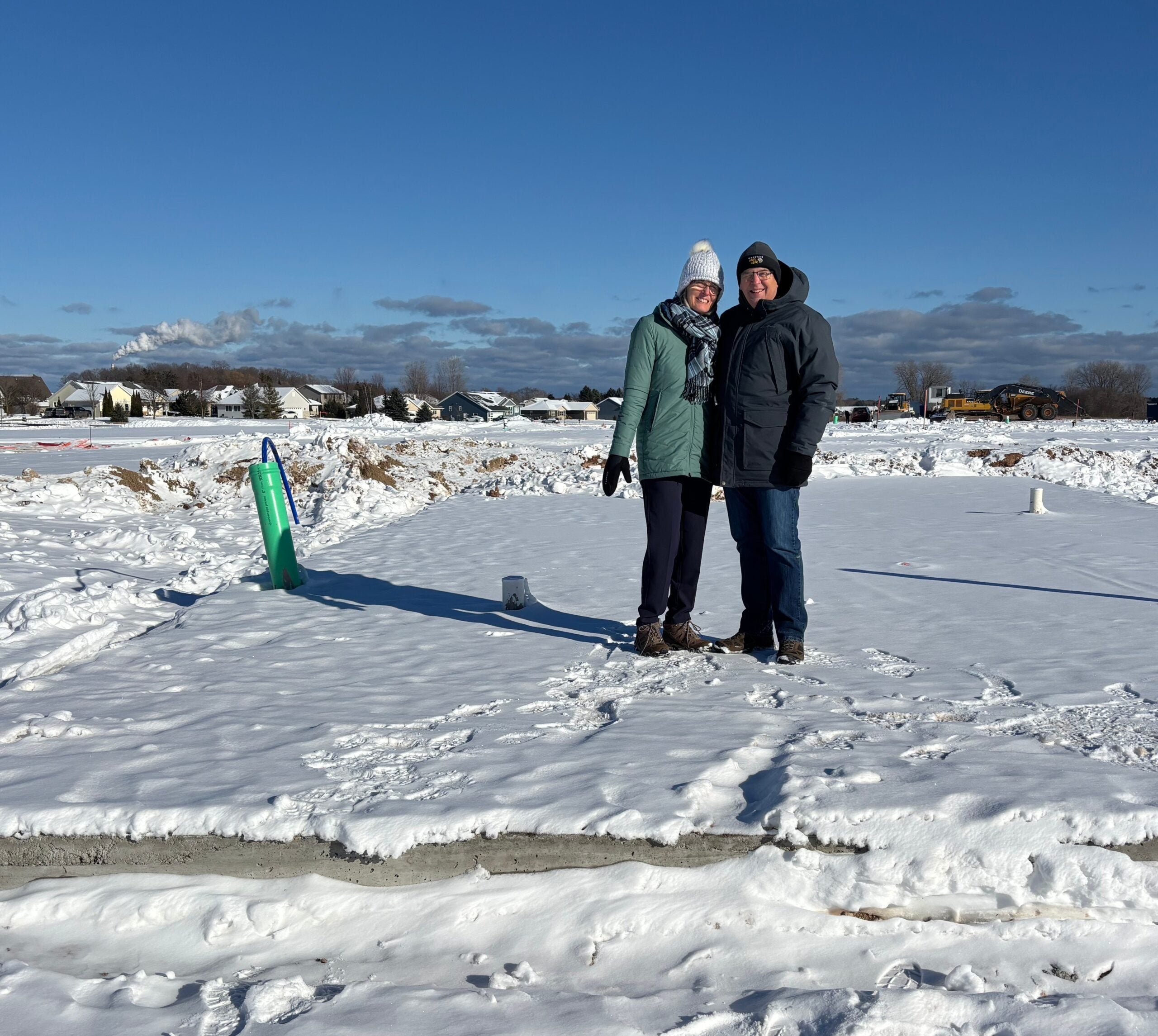 Two people wearing winter clothing stand on a snow-covered construction site, with houses, machinery, and a clear blue sky in the background.