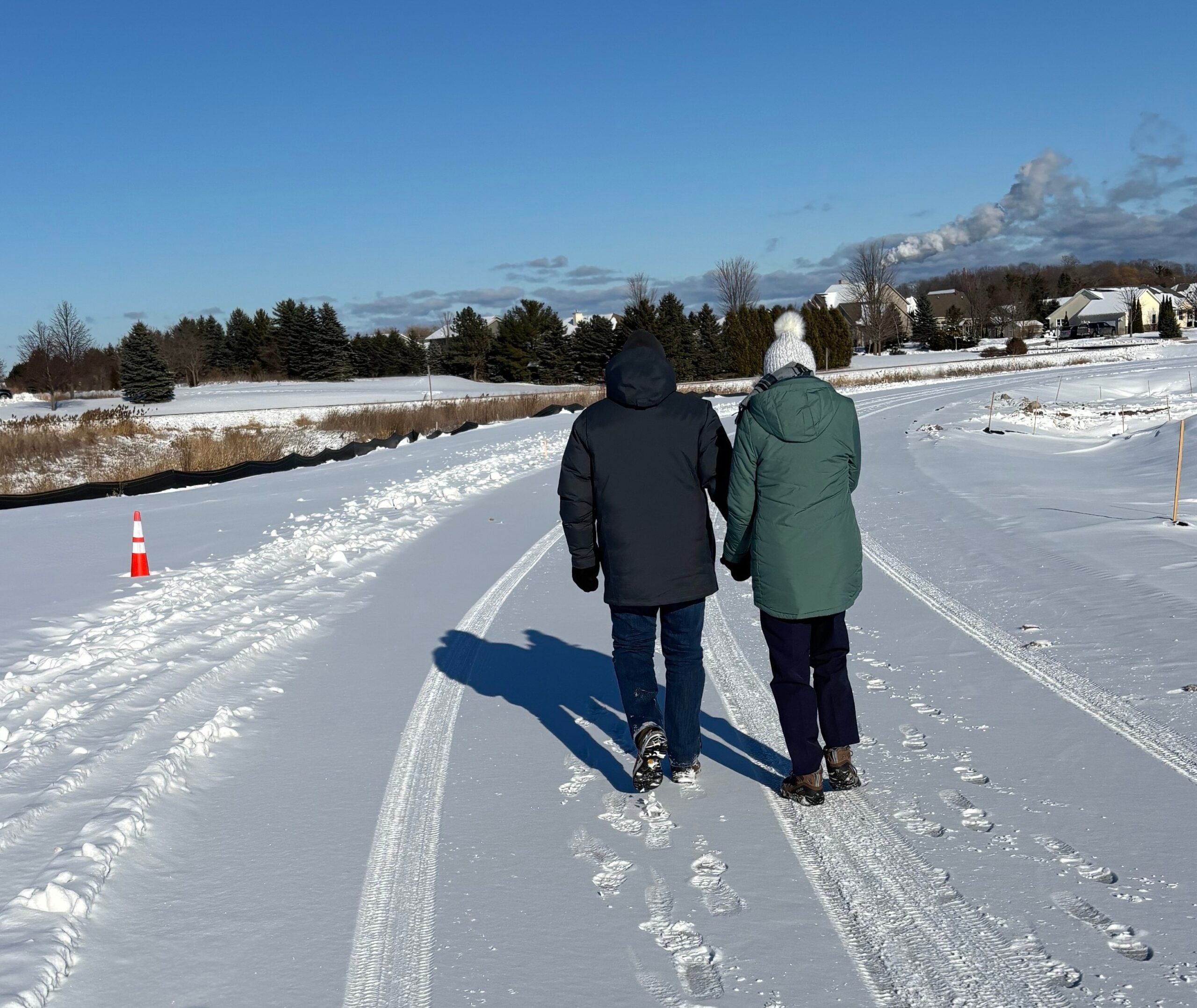 Two people walk side by side on a snow-covered path with visible tracks, surrounded by open fields and houses under a clear blue sky.