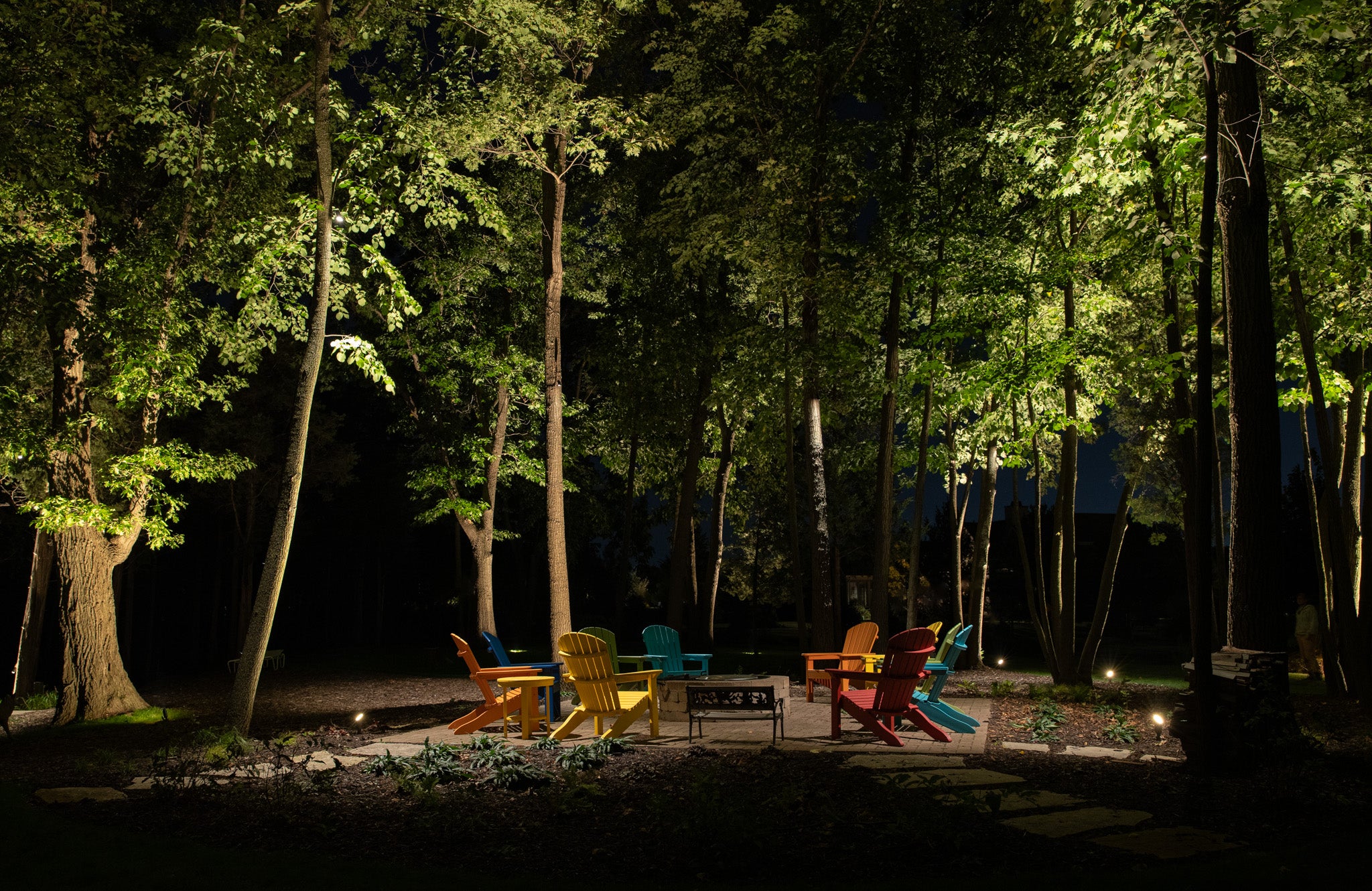 A group of colorful Adirondack chairs surrounds a small fire pit in a wooded area at night, illuminated by outdoor lighting.