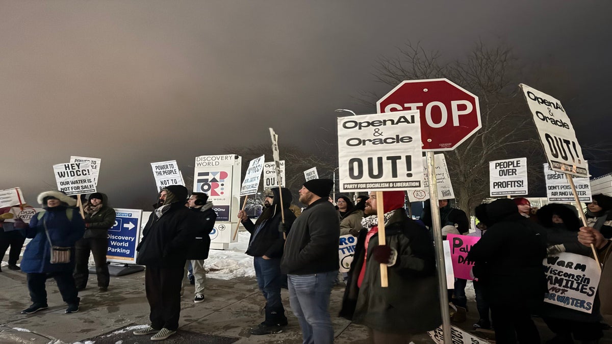 A group of people stand on a snowy sidewalk at night holding protest signs, including messages against OpenAI, Oracle, and in support of workers’ rights.