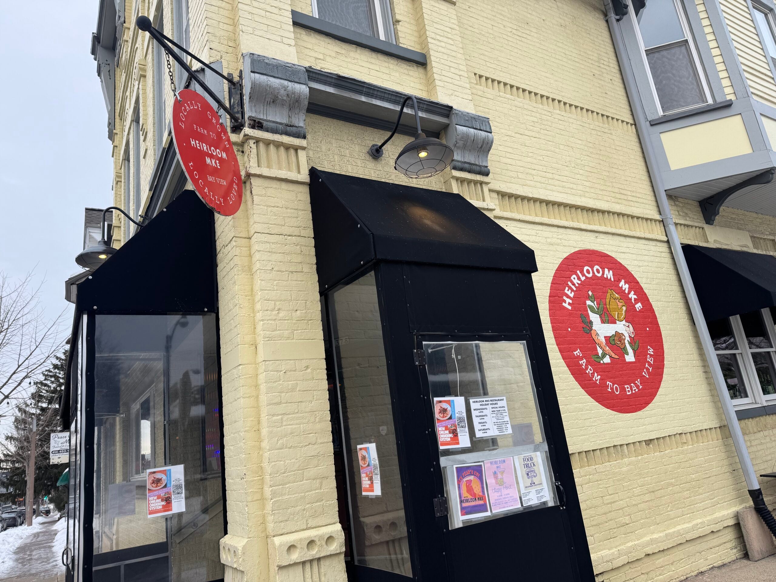 Exterior of Heirloom MX restaurant, featuring a red circular logo with a farm image and “Heirloom MX Farm to Bay Taqueria” text, and a red hanging sign on a light yellow brick building.