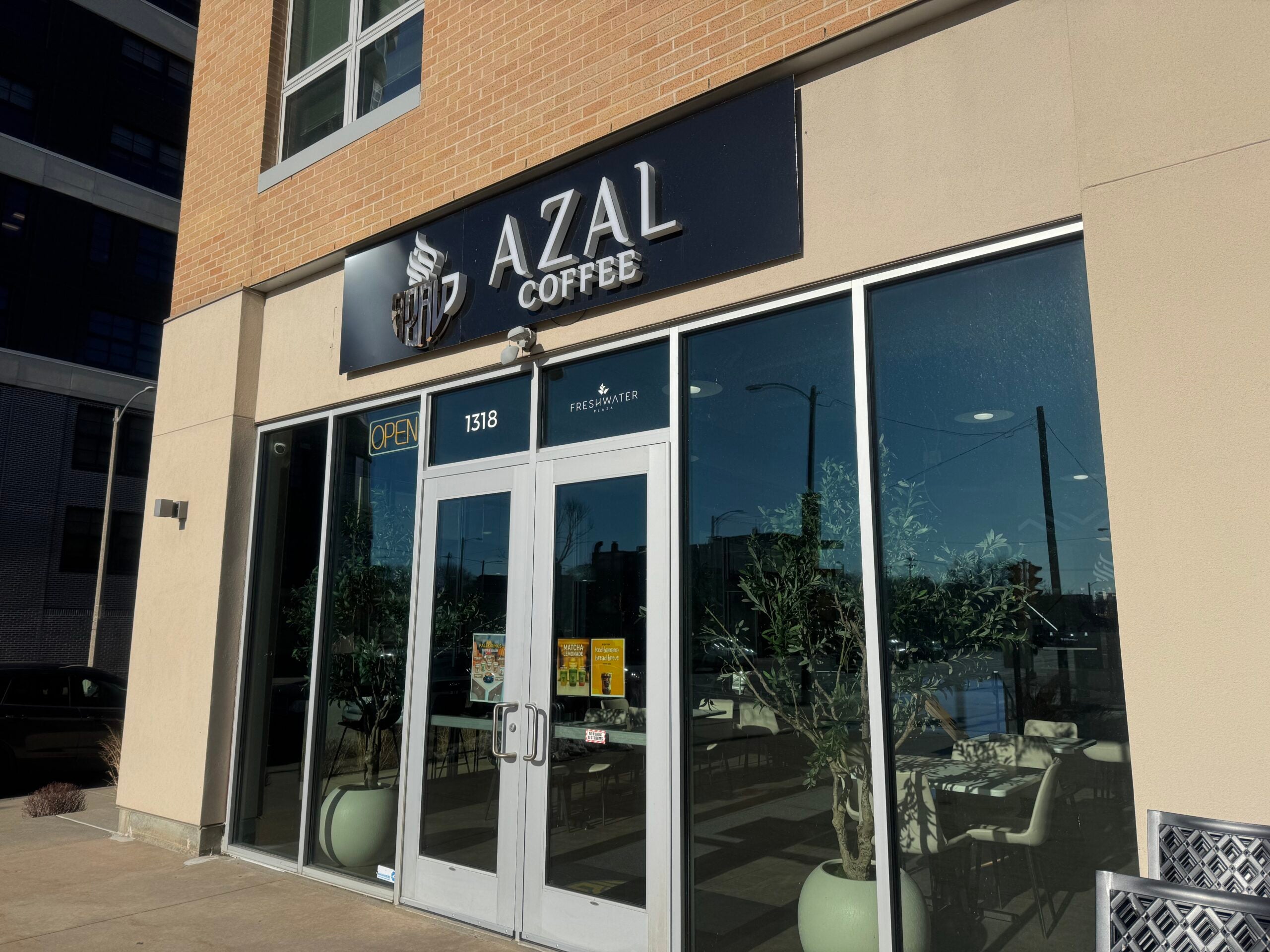 The exterior of Azal Coffee shop with glass windows, double doors, and a sign above reading AZAL COFFEE. Two potted plants are placed by the entrance.
