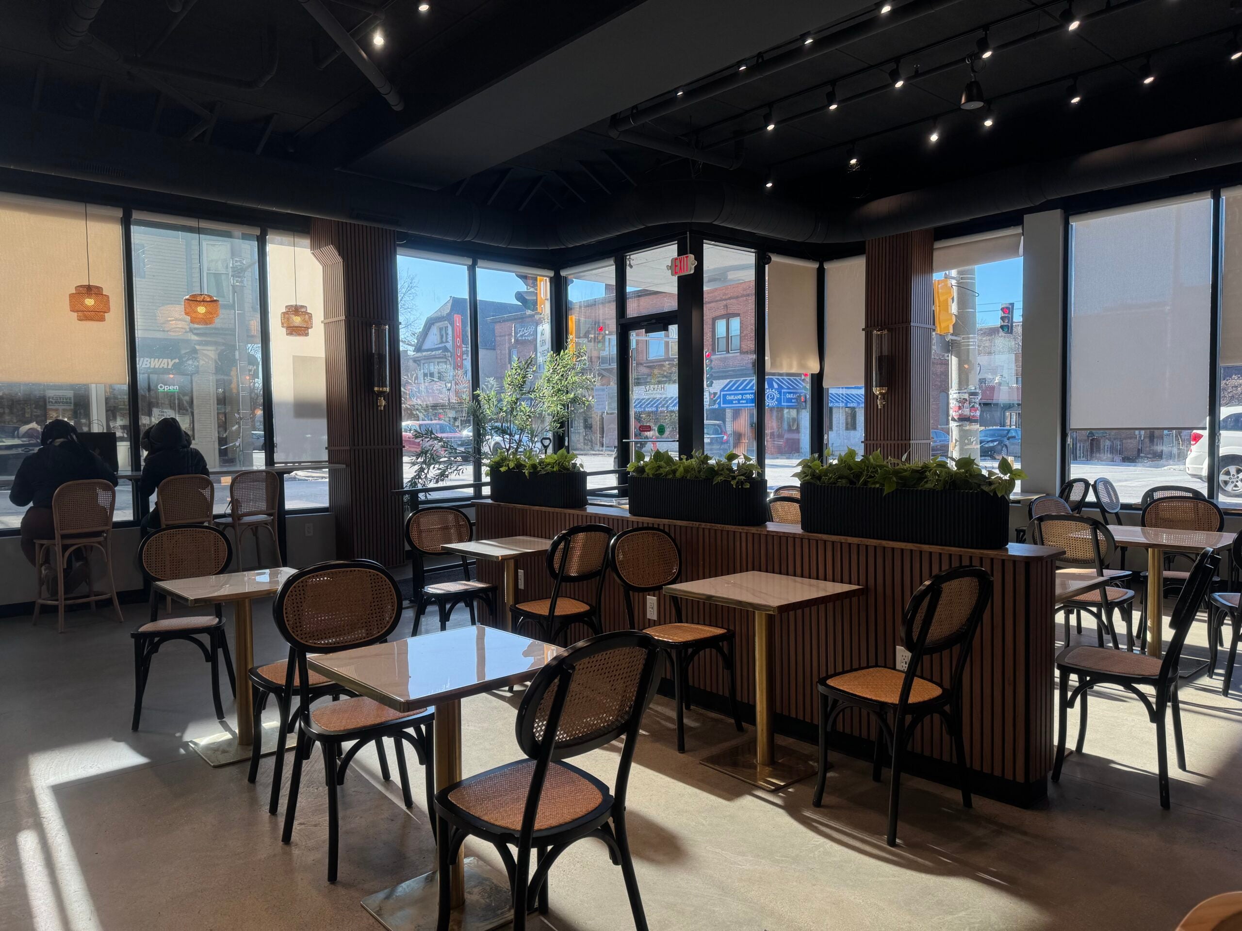 A modern café with wooden tables and chairs, large windows letting in sunlight, a few people seated, and plants dividing the seating area.