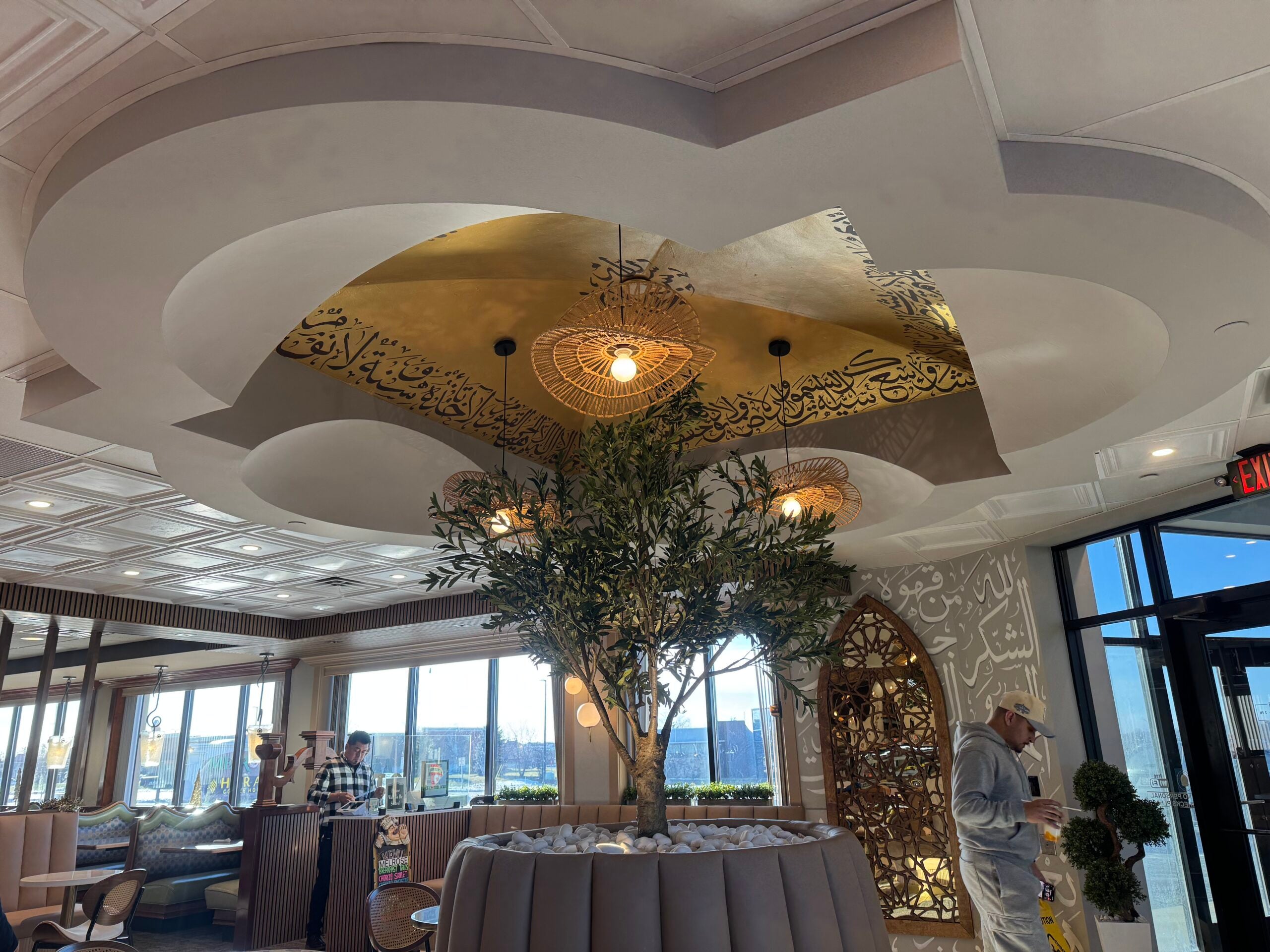 A modern restaurant interior with a central tree planter, Arabic calligraphy on the ceiling, geometric decor elements, and two people inside.