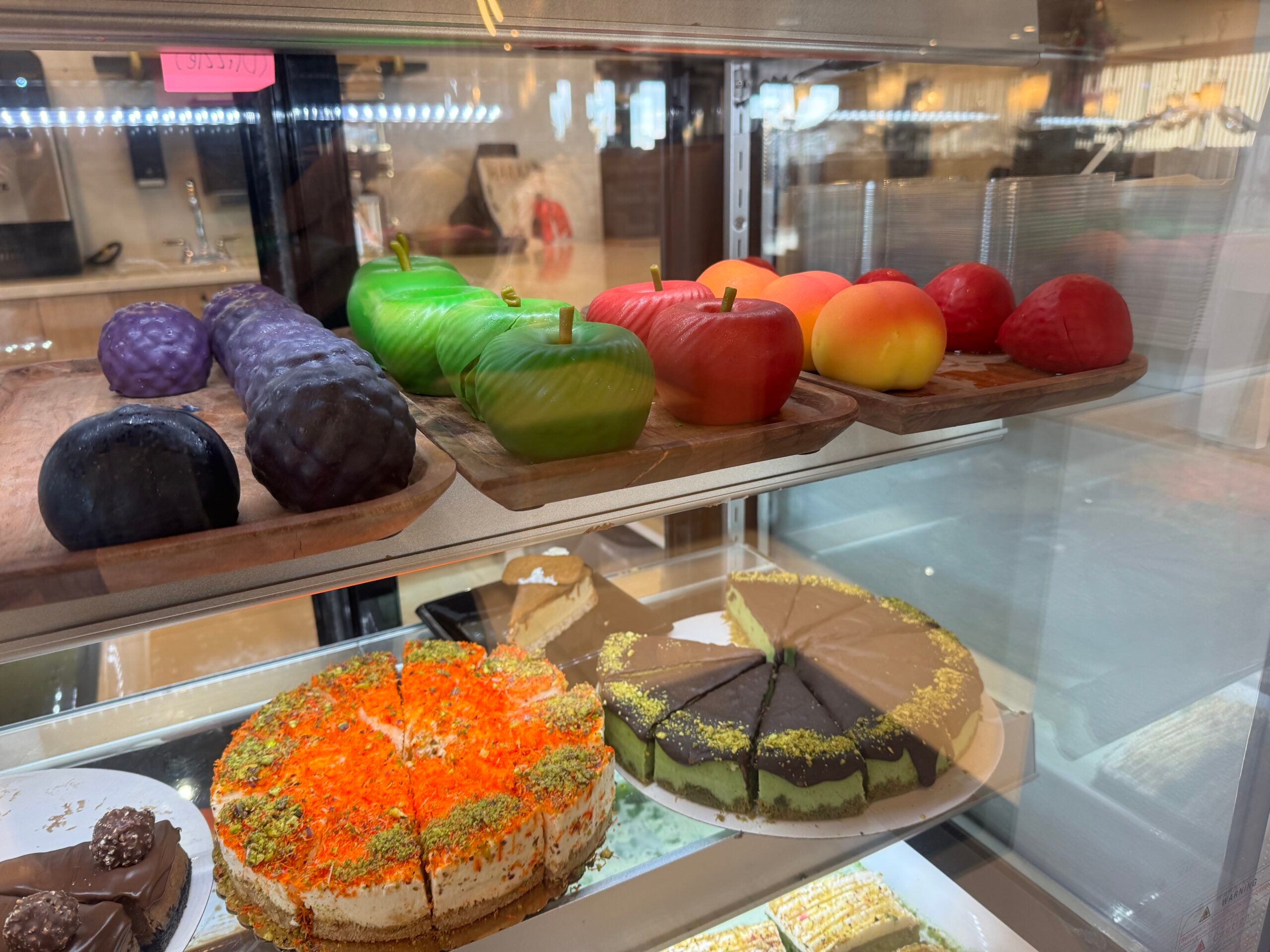 Cakes and pastries displayed in a refrigerated case, including desserts shaped like apples and grapes, and several sliced cakes on lower shelves.