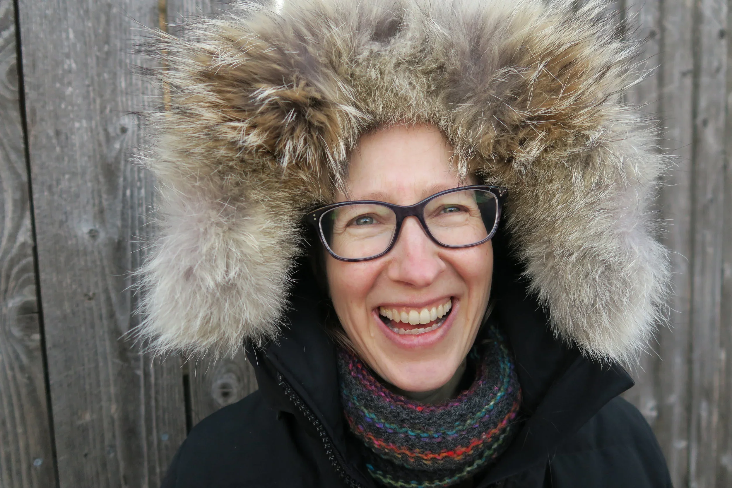 A person wearing glasses, a fur-trimmed hooded coat, and a striped scarf smiles in front of a wooden fence.