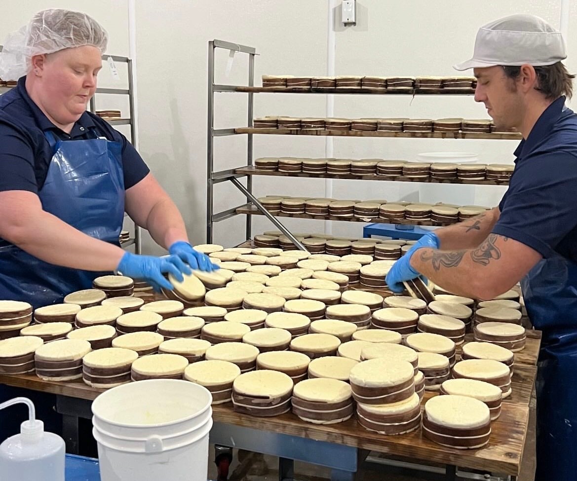 Two workers wearing gloves and hairnets assemble stacked round baked goods on a table in a commercial kitchen, with racks of finished items in the background.