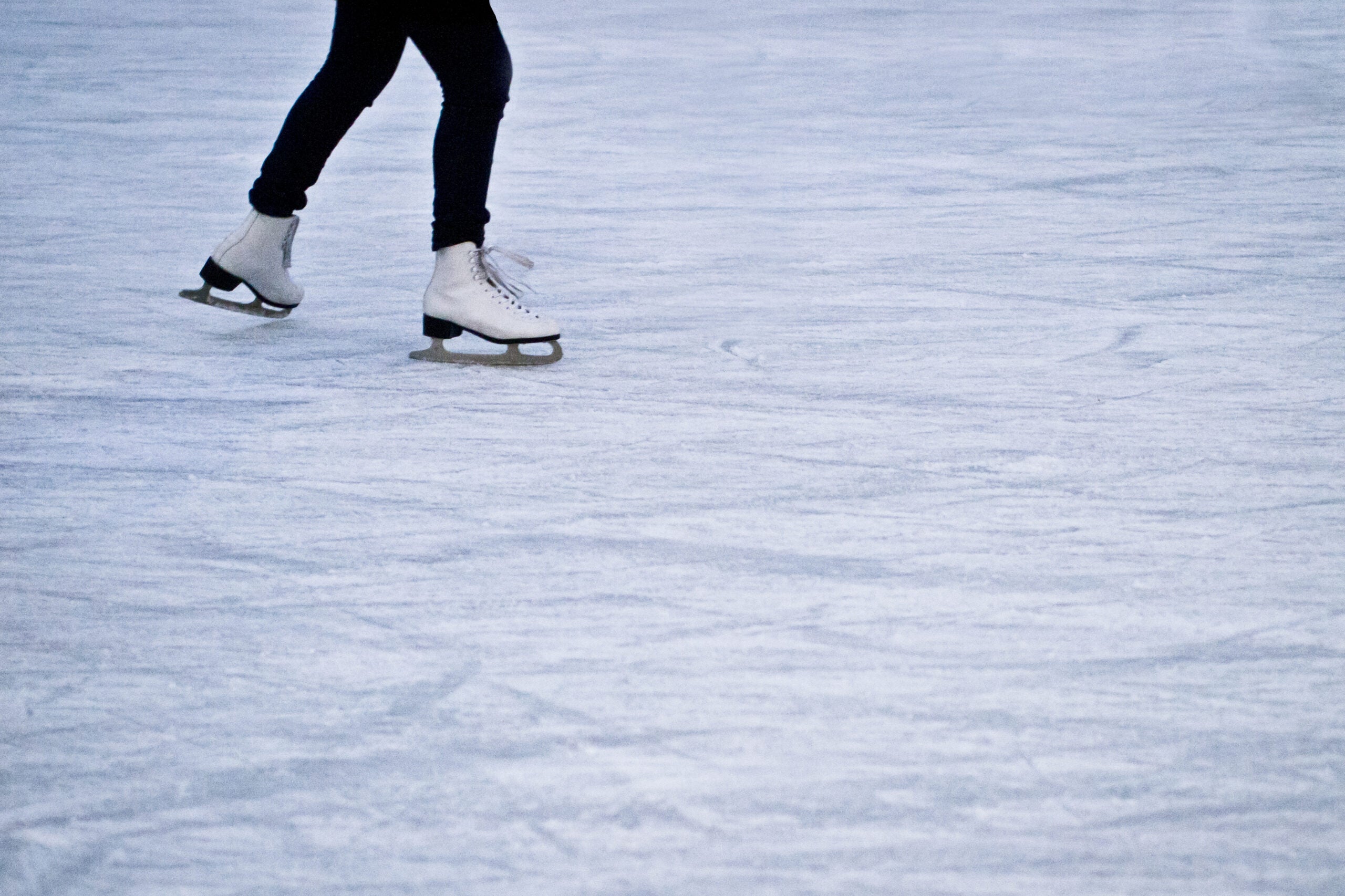 Person wearing black pants and white ice skates gliding on an ice rink. Only the legs and lower skates are visible.